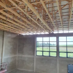 Wnętrze budynku w trakcie prac wykończeniowych: surowy tynk na ścianach, drewniana konstrukcja dachu, widok na zewnątrz przez okna z zielonym krajobrazem, podnośnik koszowy w rogu pomieszczenia.