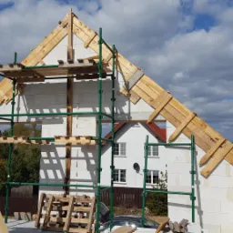 Wznoszenie ścian z białych bloczków betonu komórkowego, widoczne rusztowanie, drewniane elementy konstrukcji dachu oraz przygotowane zbrojenie na fundamencie, w tle budynek z czerwonym dachem.