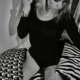 Portret kobiety w czarnym body, siedzącej na poduszkach w zebry i z wzorem kotów, z delikatnym makijażem i upiętymi włosami, trzymającej pasmo włosów w palcach, z zegarkiem na nadgarstku.