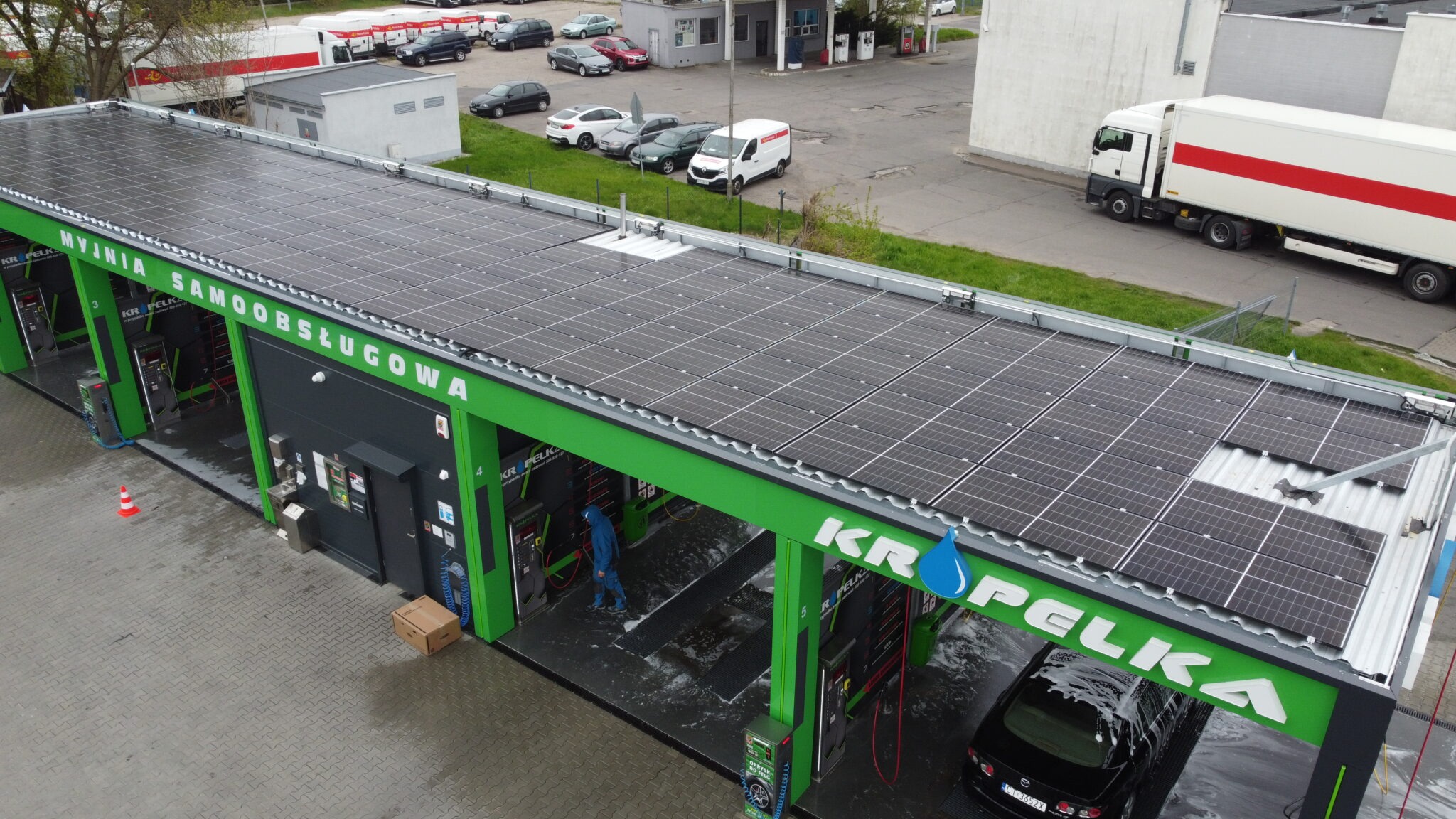 Widok z góry na samoobsługową myjnię samochodową z panelami słonecznymi na dachu, gdzie osoba w niebieskim kombinezonie czyści stanowisko, a czarny samochód jest myty w sąsiednim stanowisku.