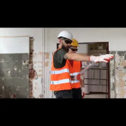 Dwóch mężczyzn w kaskach i kamizelkach odblaskowych, analizujących plan na tle częściowo zdemontowanej ściany wewnątrz budynku.