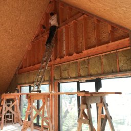 MwkBudPoland - Pracownik na drabinie montuje konstrukcję drewnianą poddasza z izolacją z wełny mineralnej, widoczne rusztowania i okna.