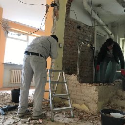 MwkBudPoland - Dwóch mężczyzn w trakcie prac rozbiórkowych ściany działowej wewnątrz budynku, widoczne odsłonięte cegły, instalacje i gruz na podłodze, drabina oparta o ścianę.
