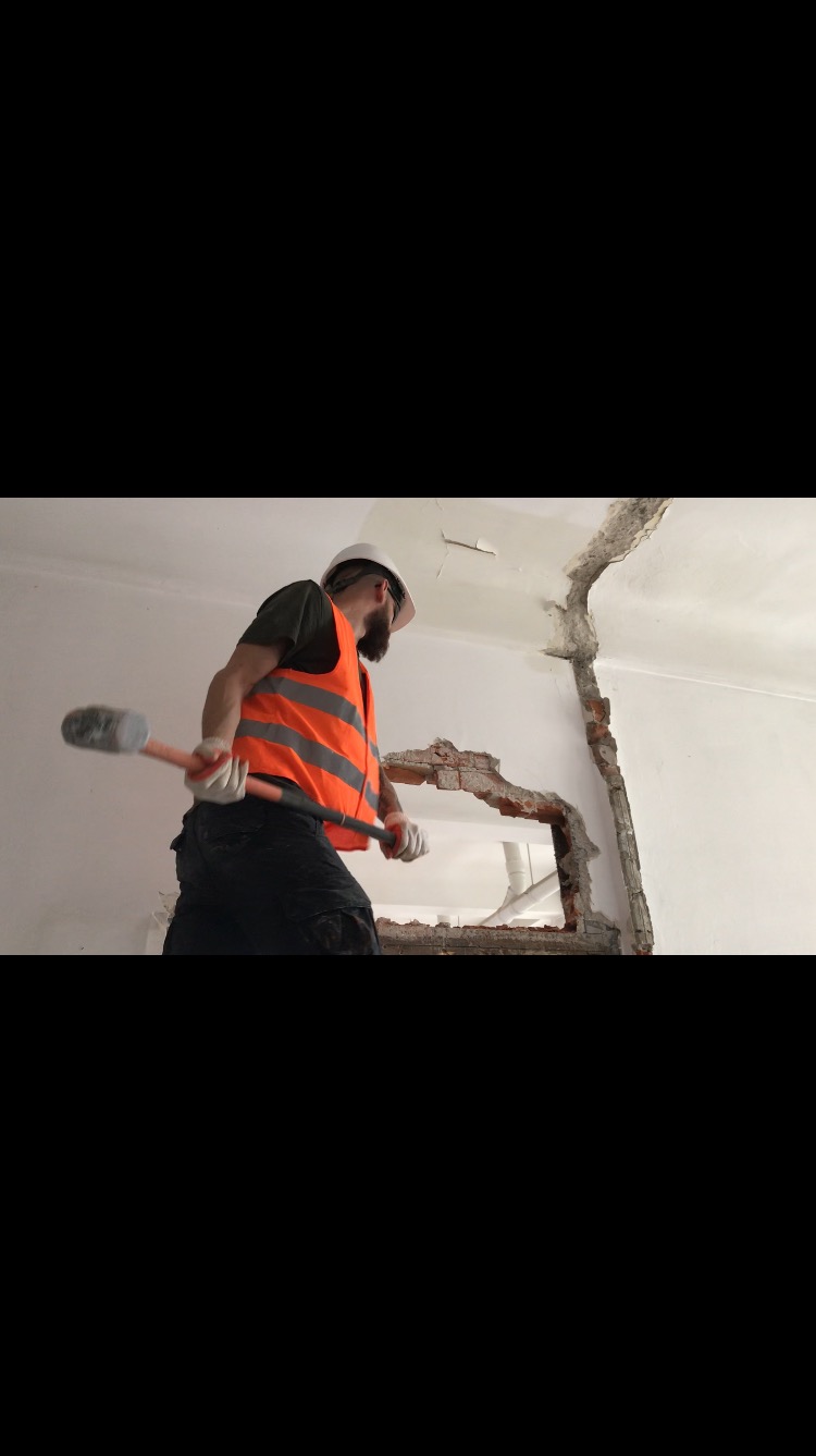 Pracownik w kasku i kamizelce odblaskowej wyburza fragment ściany młotem, widoczne fragmenty cegieł i instalacji wewnątrz ściany.