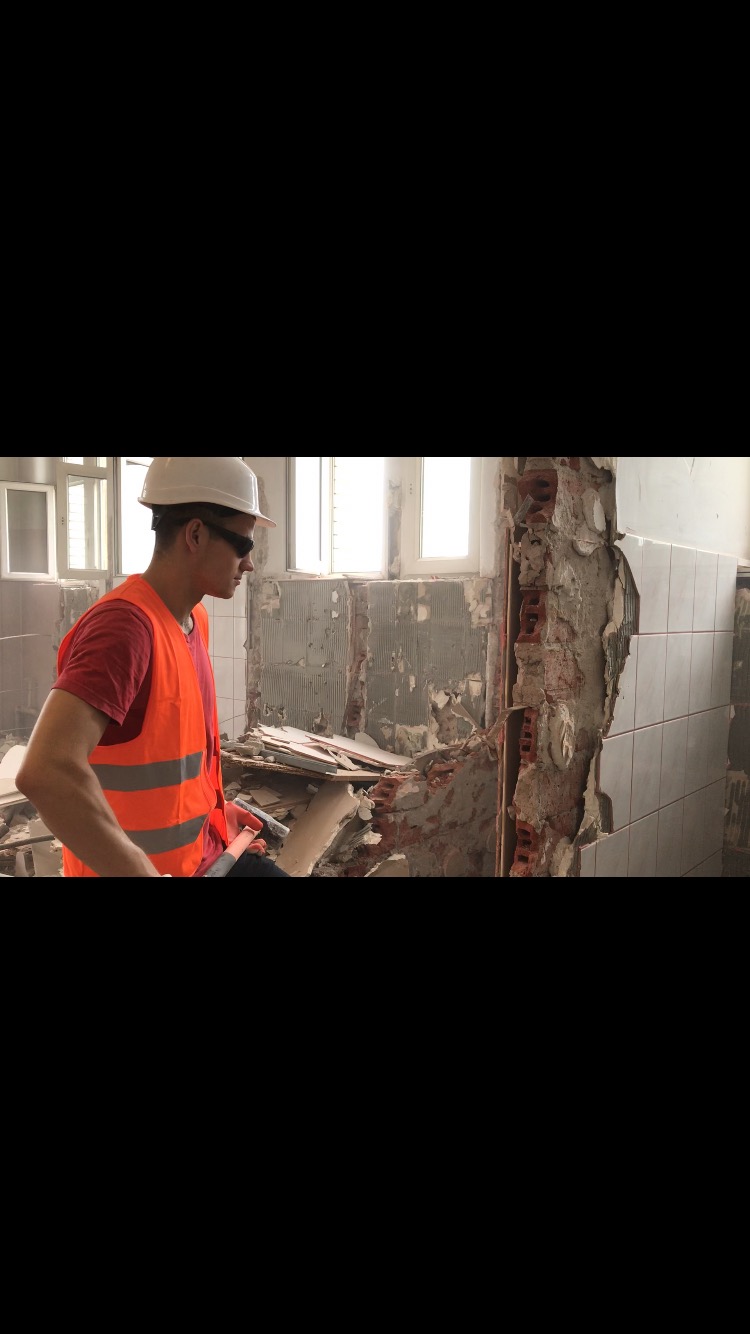 Pracownik w kasku i kamizelce odblaskowej usuwa stare płytki z fragmentu ściany w trakcie remontu łazienki, widoczne gruz i odsłonięta cegła.