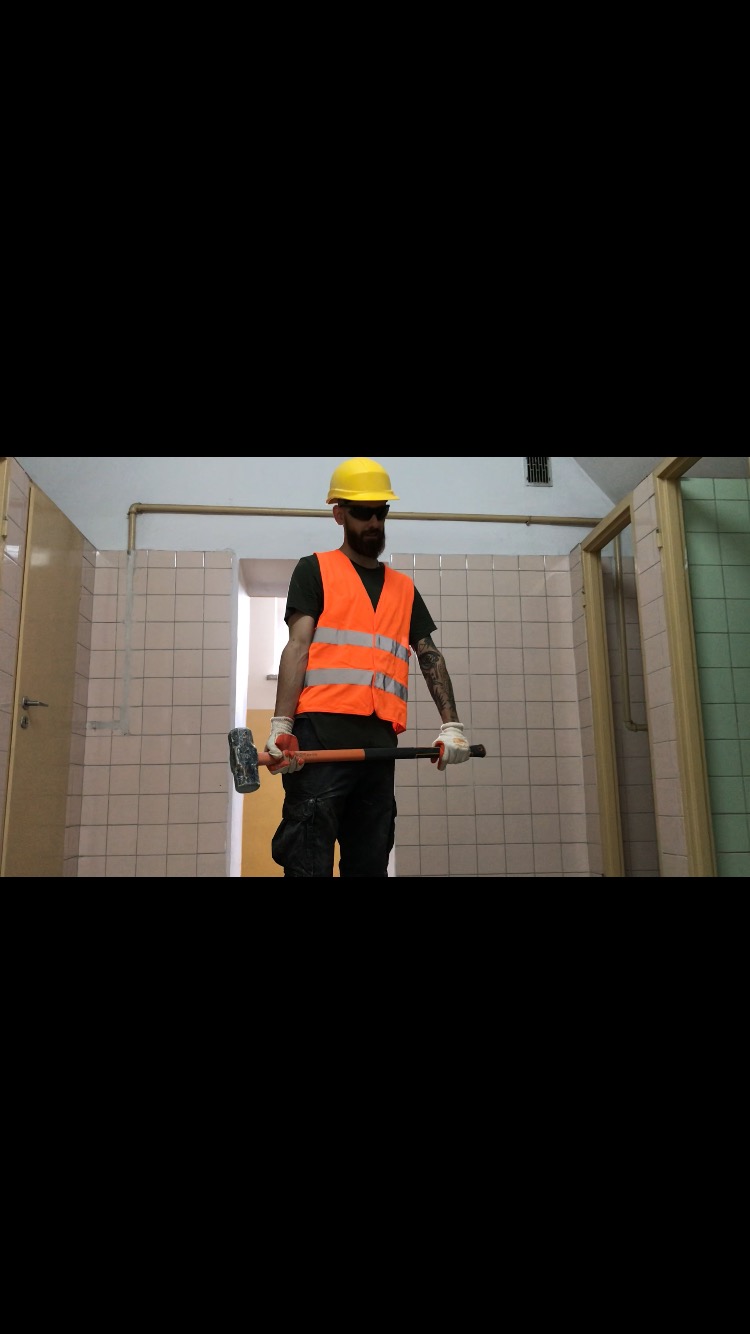 Pracownik w żółtym kasku i pomarańczowej kamizelce trzyma młot wyburzeniowy w łazience z częściowo usuniętymi płytkami, przygotowując się do dalszego demontażu.