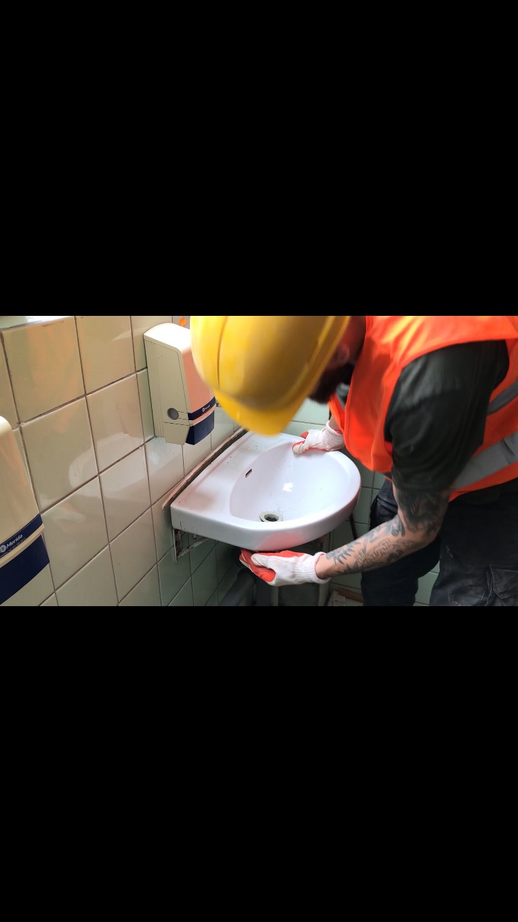 Instalator w żółtym kasku i pomarańczowej kamizelce montuje białą, narożną umywalkę w łazience z jasnymi, kwadratowymi płytkami. Widoczne rękawice robocze i tatuaże na rękach.