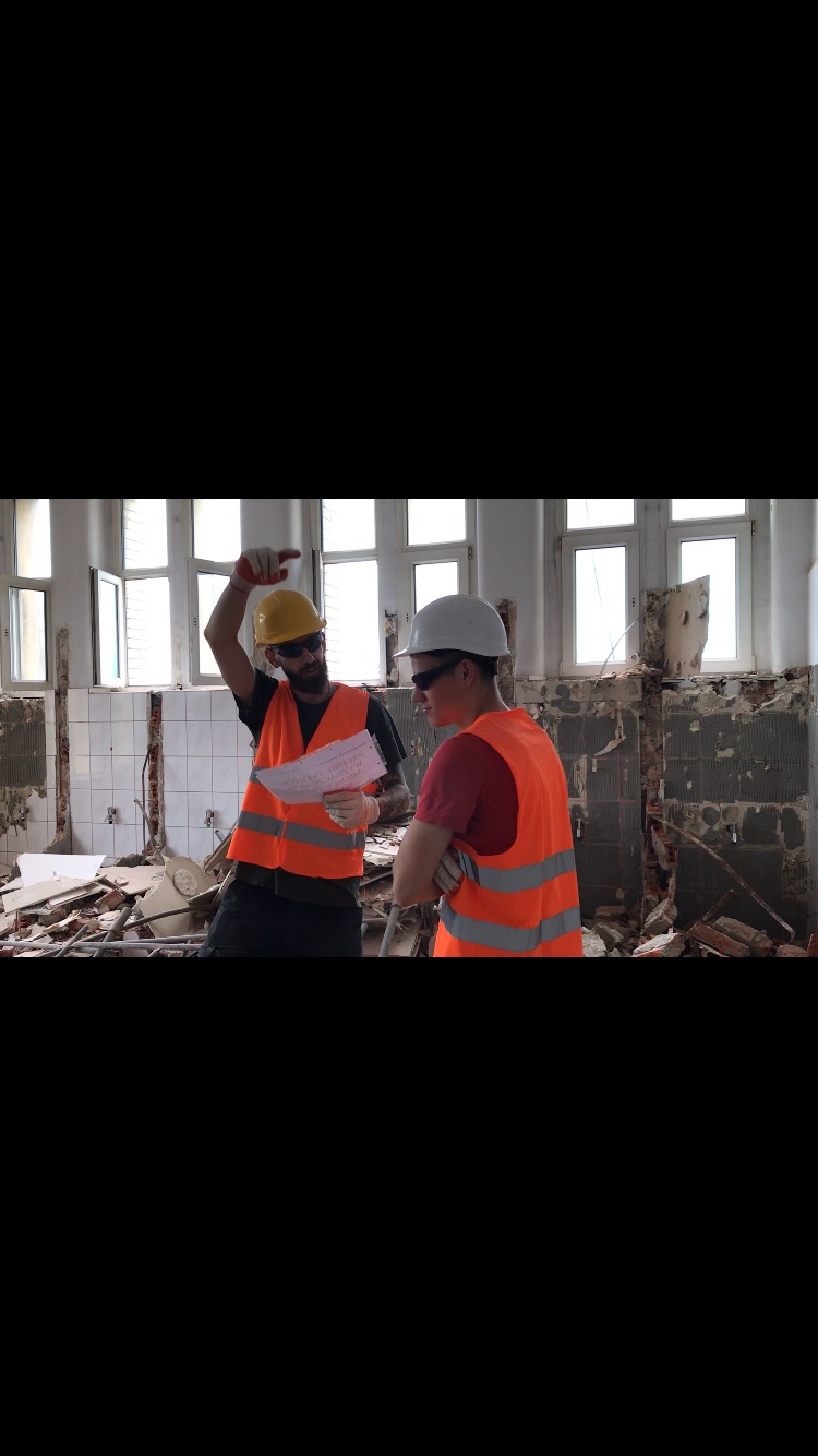 Dwóch mężczyzn w kaskach i kamizelkach odblaskowych omawia plan na tle zdemolowanego pomieszczenia z widocznymi fragmentami gruzu i uszkodzonych ścian.