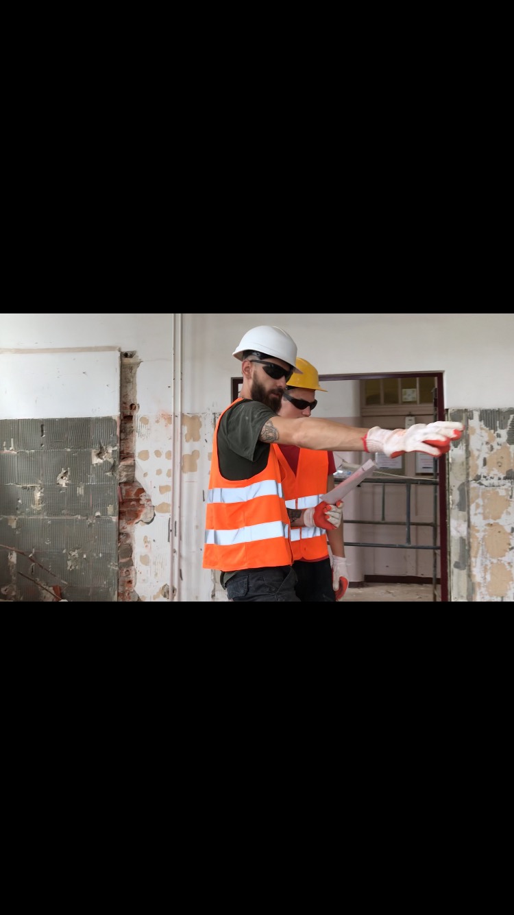 Dwóch mężczyzn w kaskach i kamizelkach odblaskowych, analizujących plan na tle częściowo zdemontowanej ściany wewnątrz budynku.