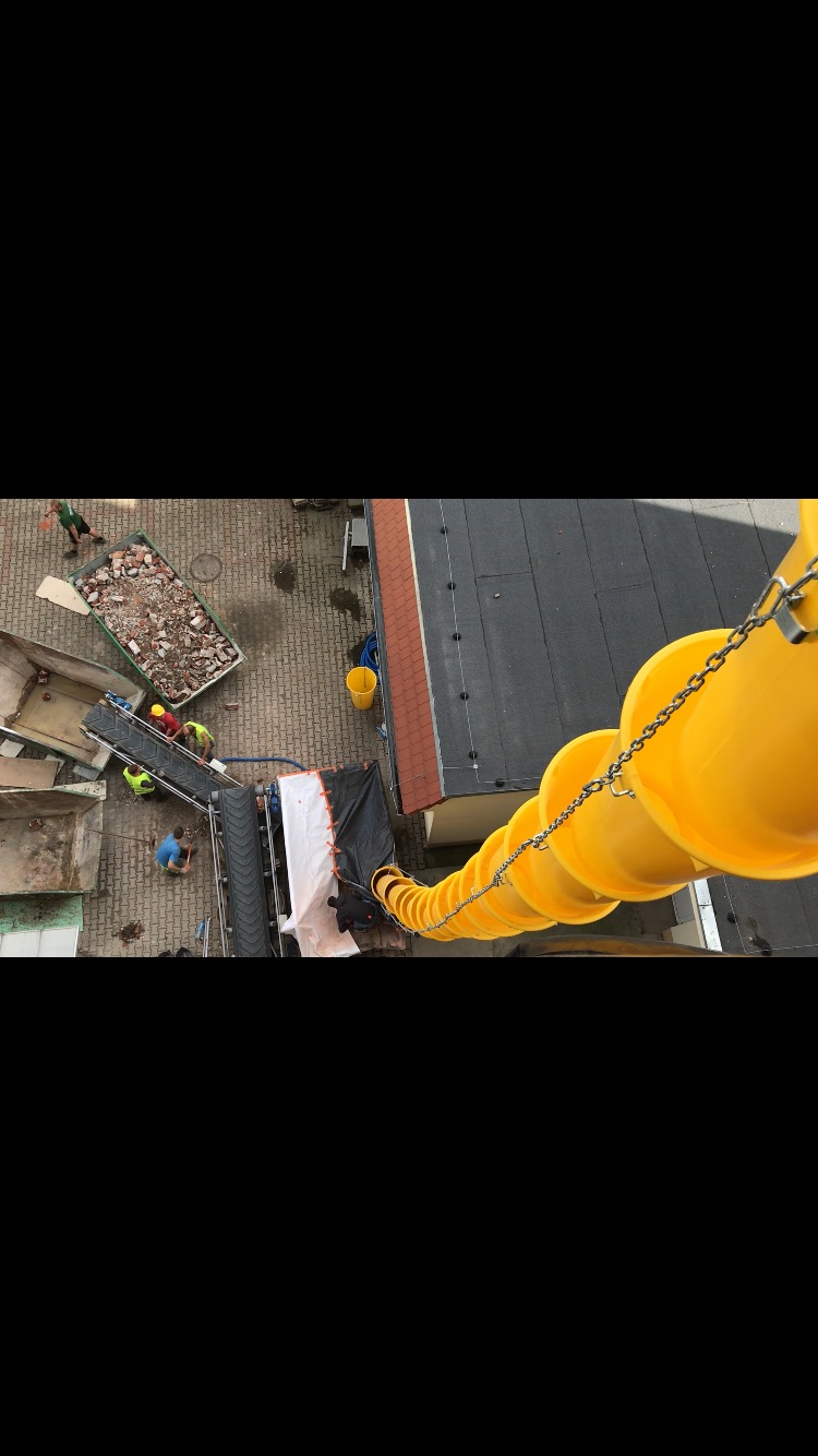 Widok z góry na plac budowy z żółtą rurą zsypową, kontenerem wypełnionym gruzem, taśmociągiem i pracownikami w kamizelkach odblaskowych.