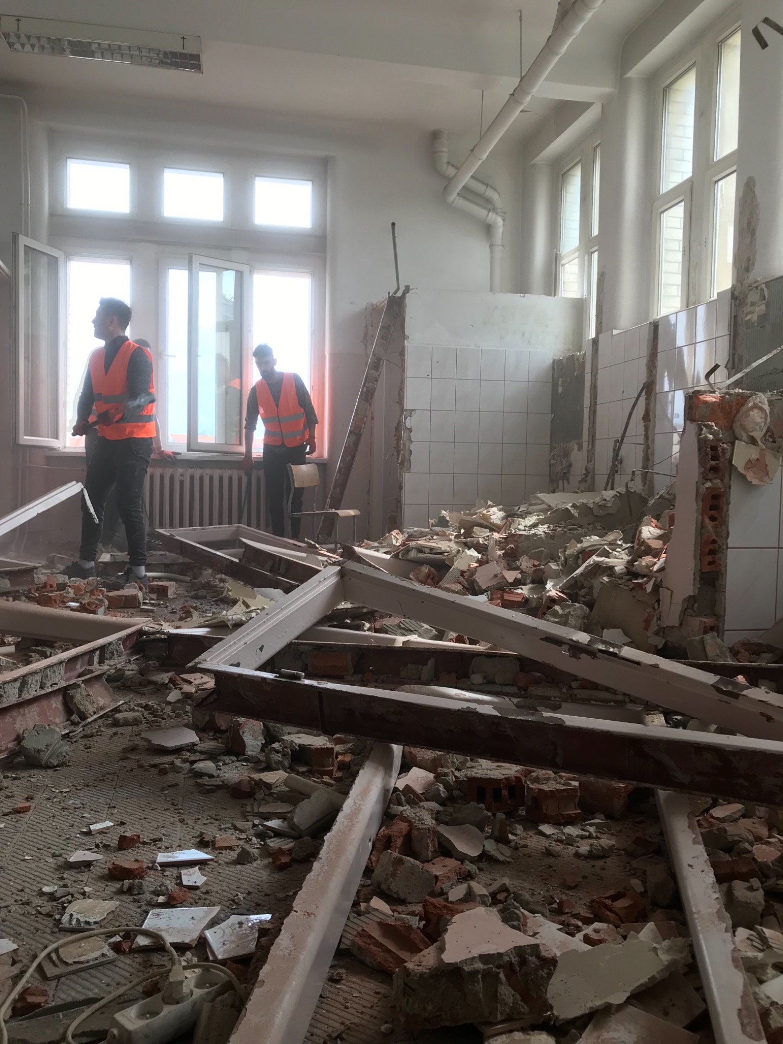 Wnętrze w trakcie demontażu: dwóch pracowników w kamizelkach odblaskowych stoi przy oknie, a na podłodze gruz, cegły i fragmenty ścian.