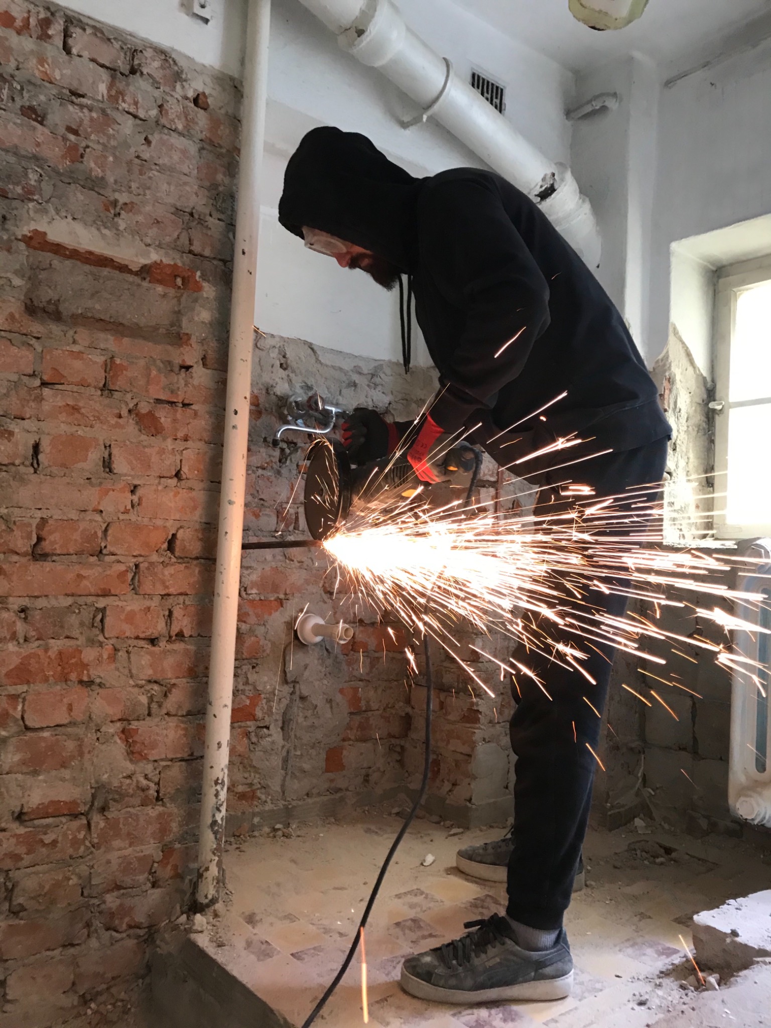 Pracownik w czarnym ubraniu i ochronnych okularach tnie metalową rurę szlifierką kątową, iskry rozbryzgują się na tle ceglanej ściany i fragmentu okna w remontowanym pomieszczeniu.
