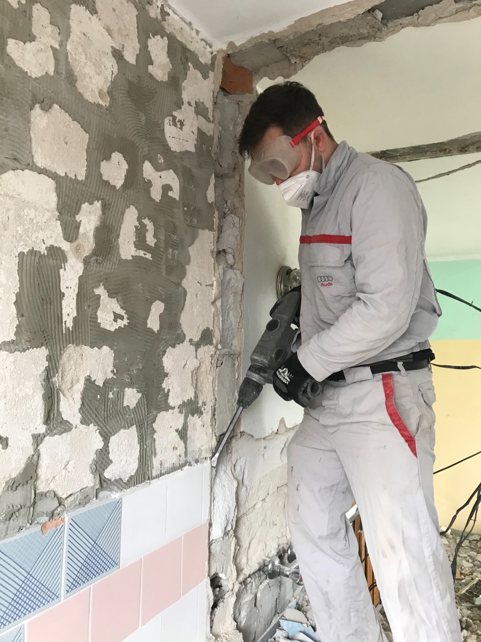 Pracownik w kombinezonie Audi, okularach ochronnych i masce usuwa stare płytki ze ściany za pomocą młota udarowego, widoczne pozostałości kleju i fragmenty płytek.