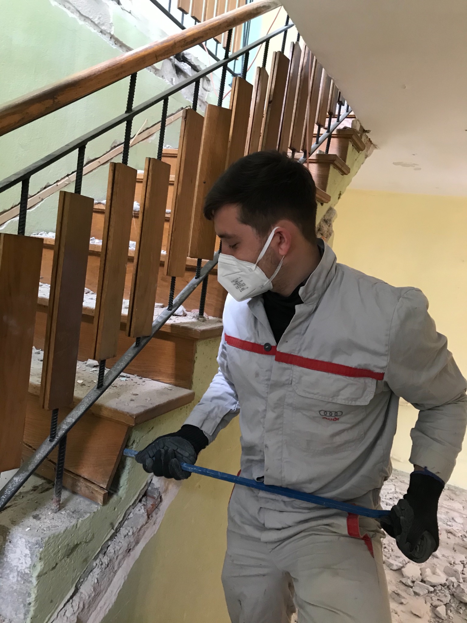 Pracownik w kombinezonie z logo Audi, w masce ochronnej, używa łomu do demontażu schodów z drewnianymi balustradami, widoczne uszkodzenia ściany i gruz.