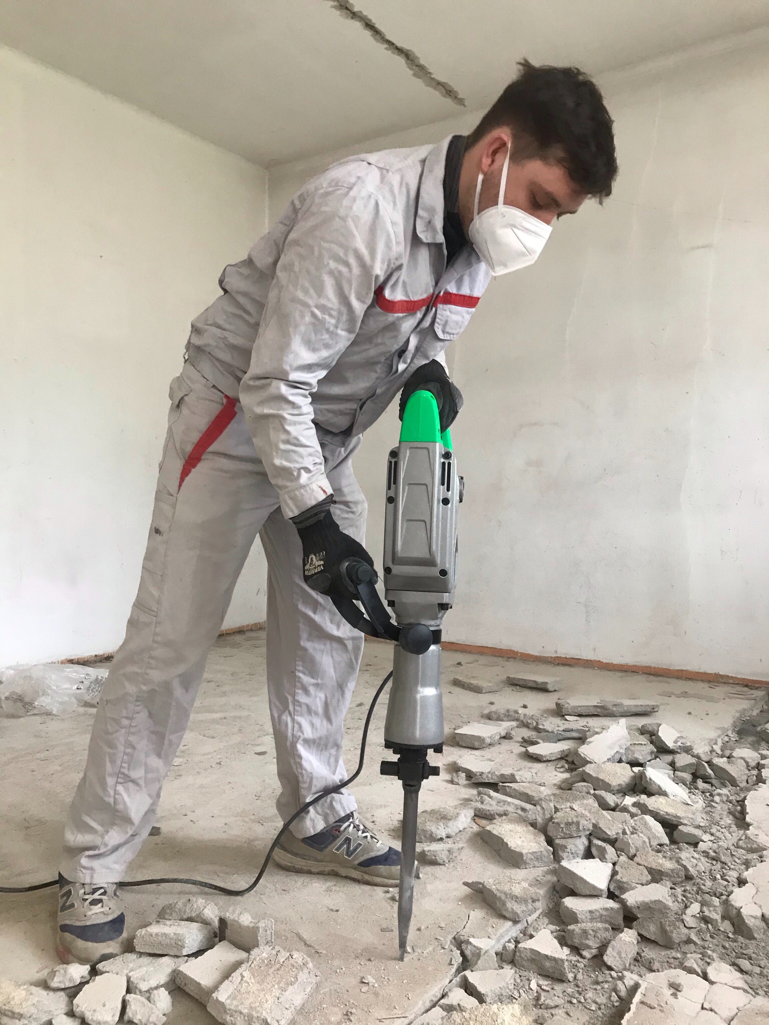 Pracownik w kombinezonie i masce ochronnej używa młota pneumatycznego do skuwania posadzki w pomieszczeniu w trakcie remontu.