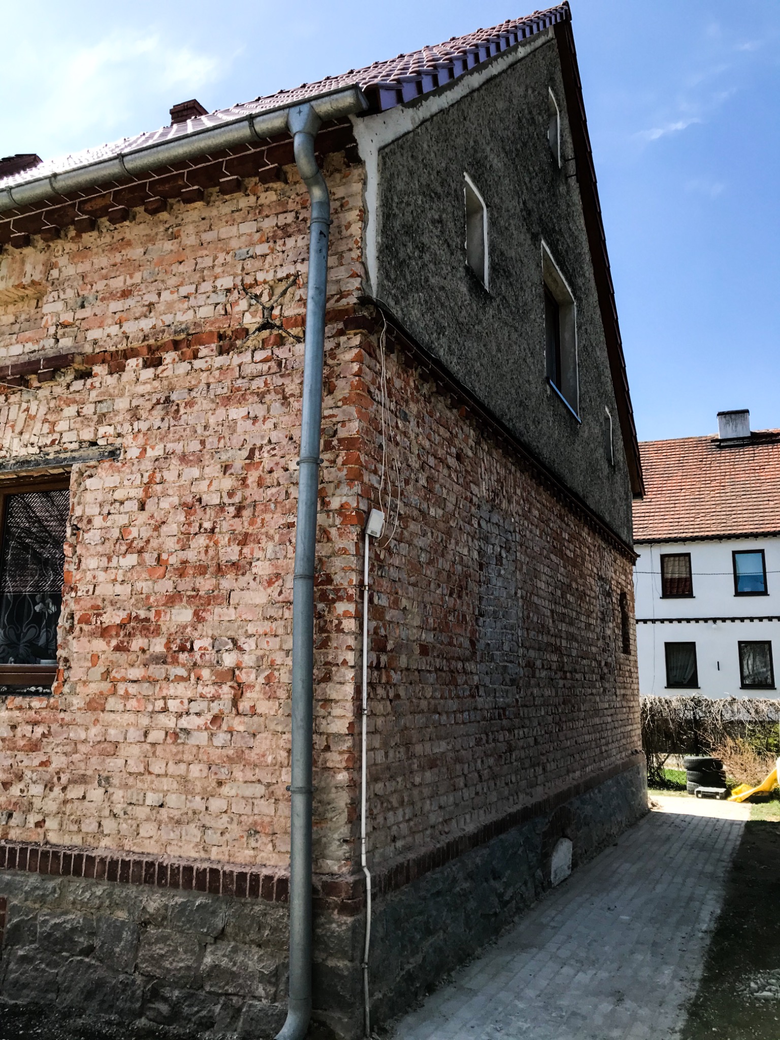 Fragment ściany budynku z czerwonej cegły, z widocznym orynnowaniem i fragmentem otynkowanej elewacji, ukazujący stan przed potencjalnym remontem lub odnowieniem.