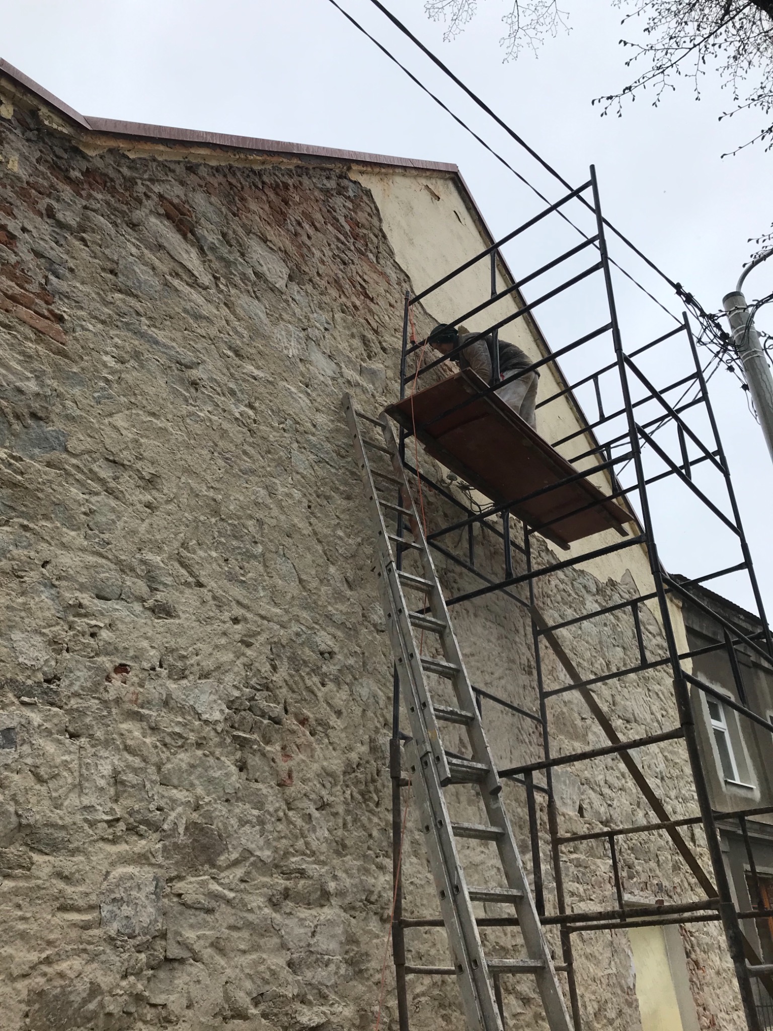 Pracownik na rusztowaniu przy kamiennej ścianie budynku, przygotowujący powierzchnię pod prace wykończeniowe, widoczna drabina i fragment okna sąsiedniego budynku.