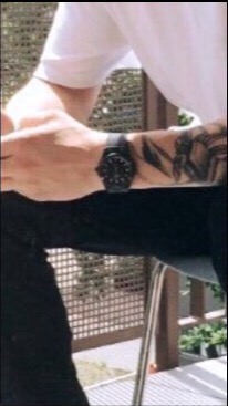 Mężczyzna w białej koszulce i czarnych spodniach siedzi na krześle. Na jego lewym nadgarstku widoczny jest czarny zegarek, a na przedramieniu tatuaż.