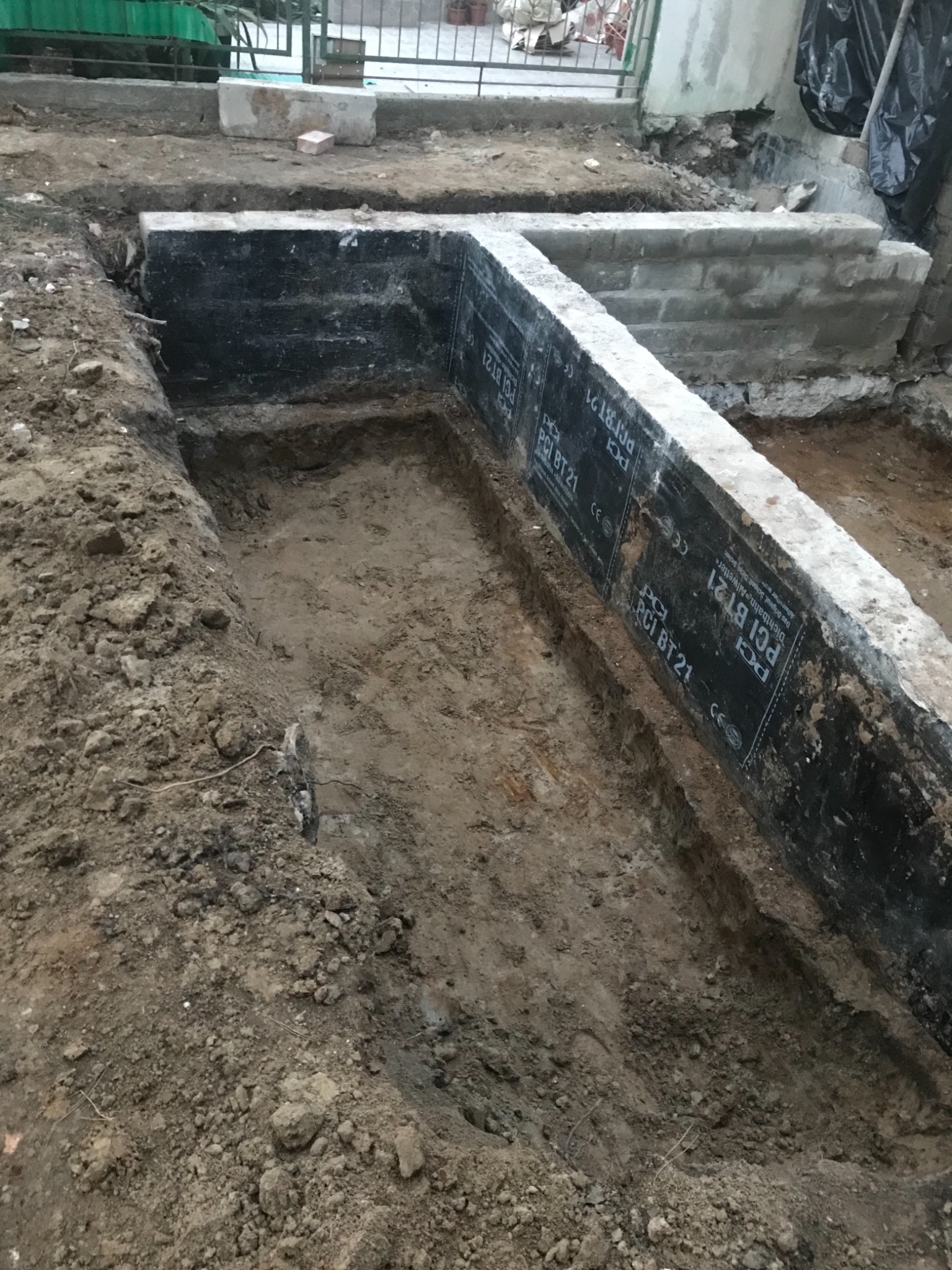Wykop fundamentowy z izolacją przeciwwilgociową PCI BT21 przy ścianie budynku, widoczne ślady prac ziemnych i betonowe elementy konstrukcyjne.