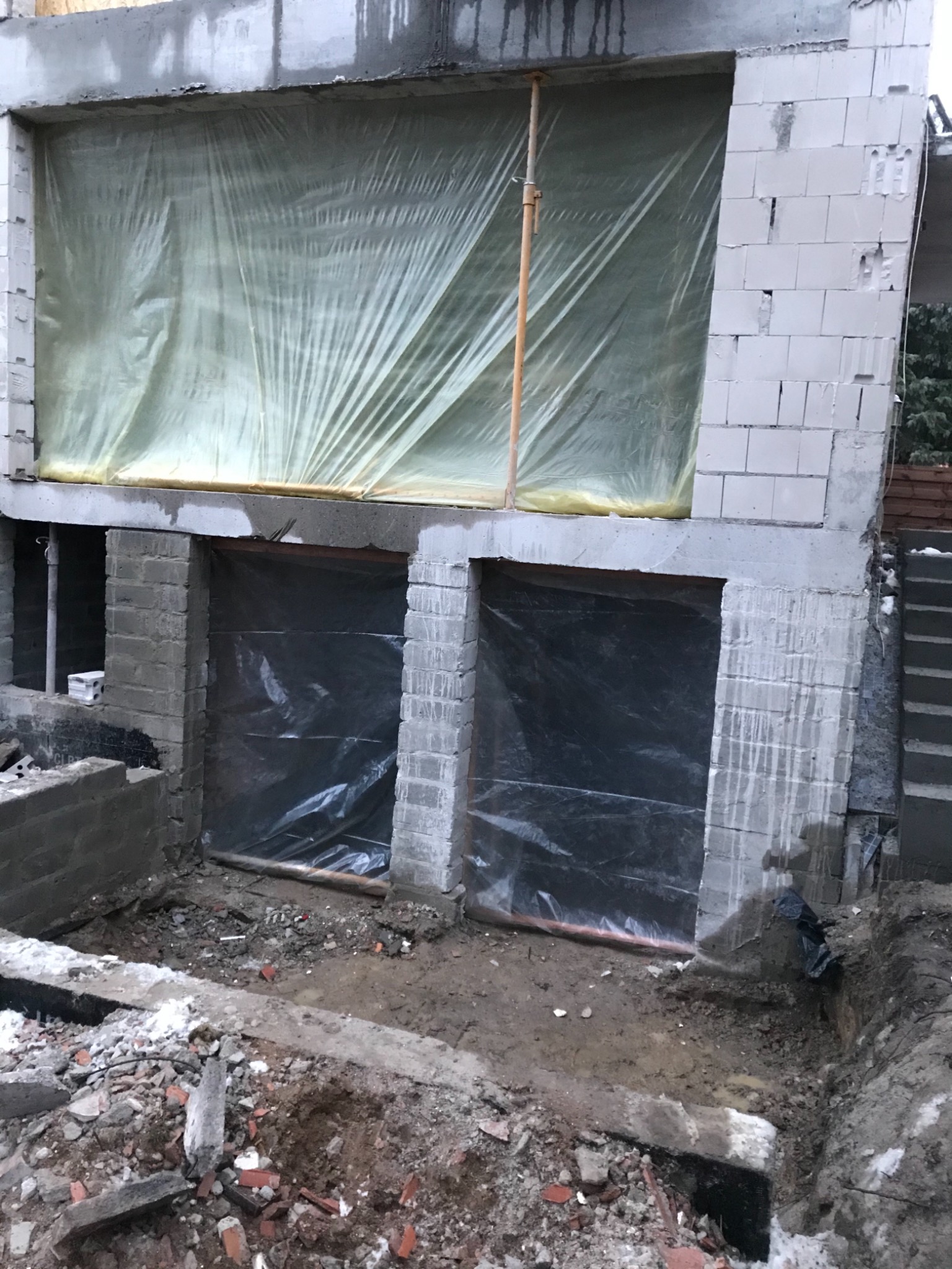 Surowy stan fundamentów budynku z pustaków betonowych, zabezpieczonych folią, z widocznym wykopem i gruzem.