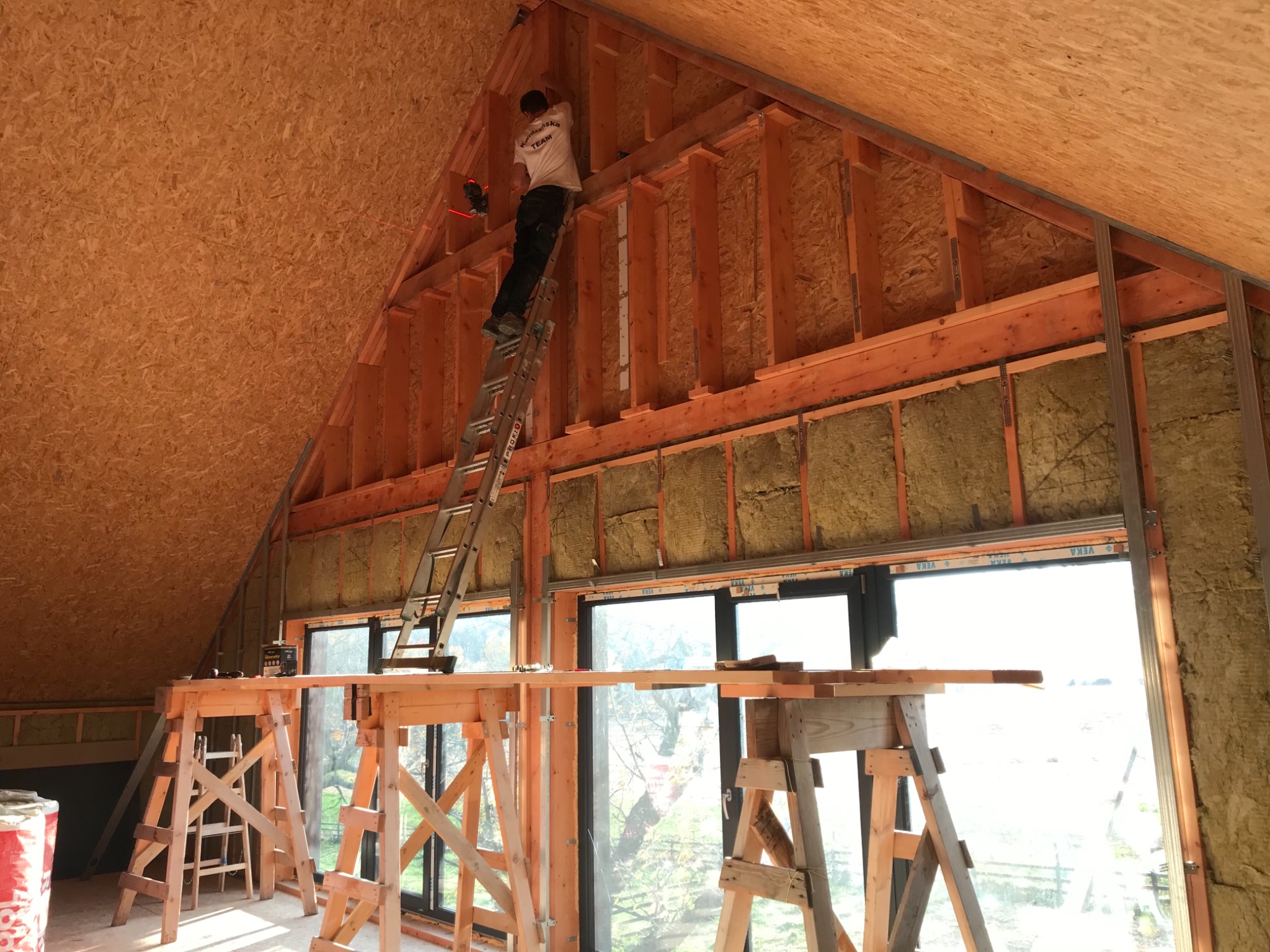 Pracownik na drabinie montuje konstrukcję drewnianą poddasza z izolacją z wełny mineralnej, widoczne rusztowania i okna.