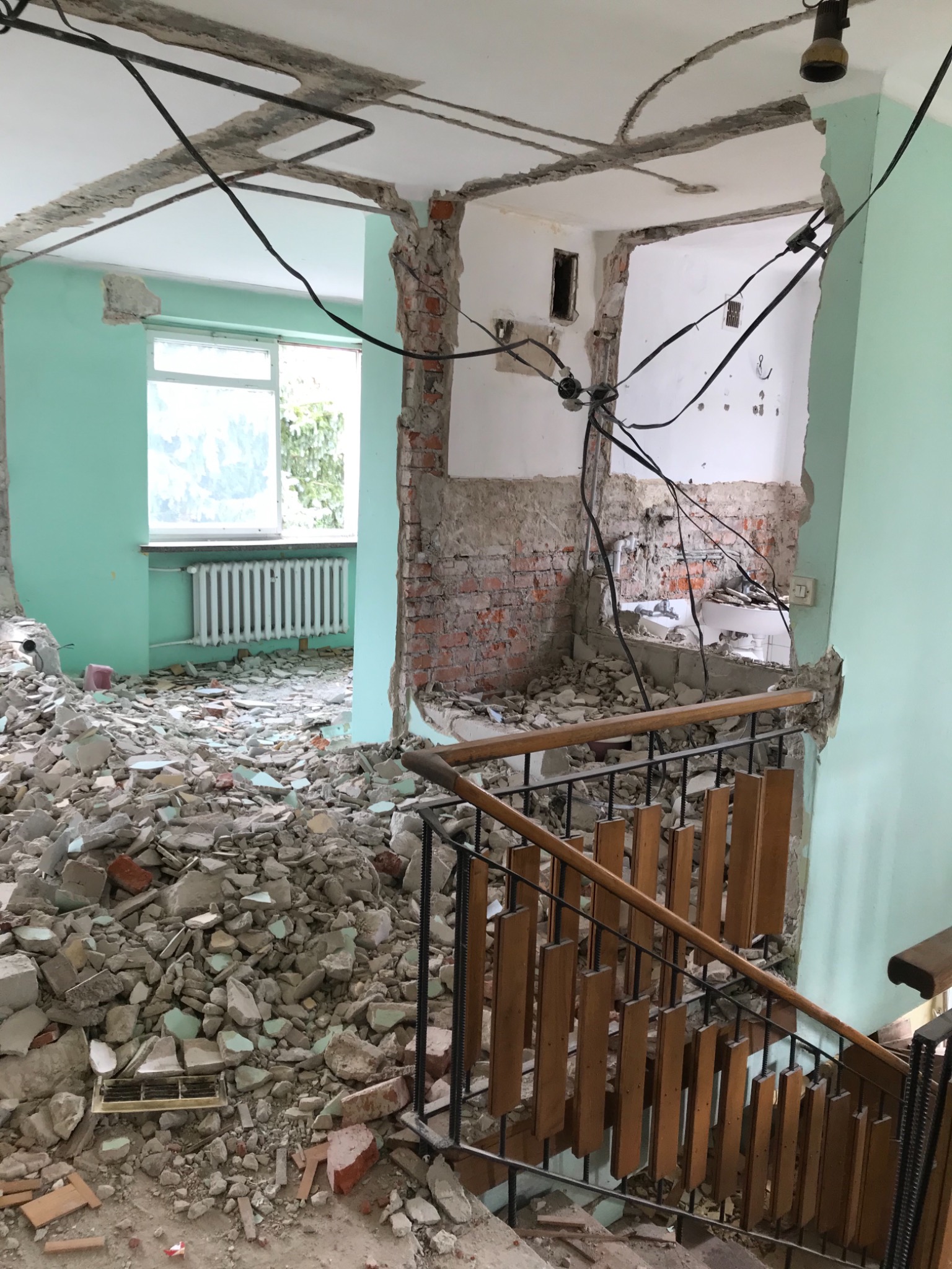 Wnętrze budynku w trakcie rozbiórki: gruz na podłodze, odsłonięte cegły, wiszące kable elektryczne, fragment schodów z drewnianą balustradą, ściany w kolorze miętowym.