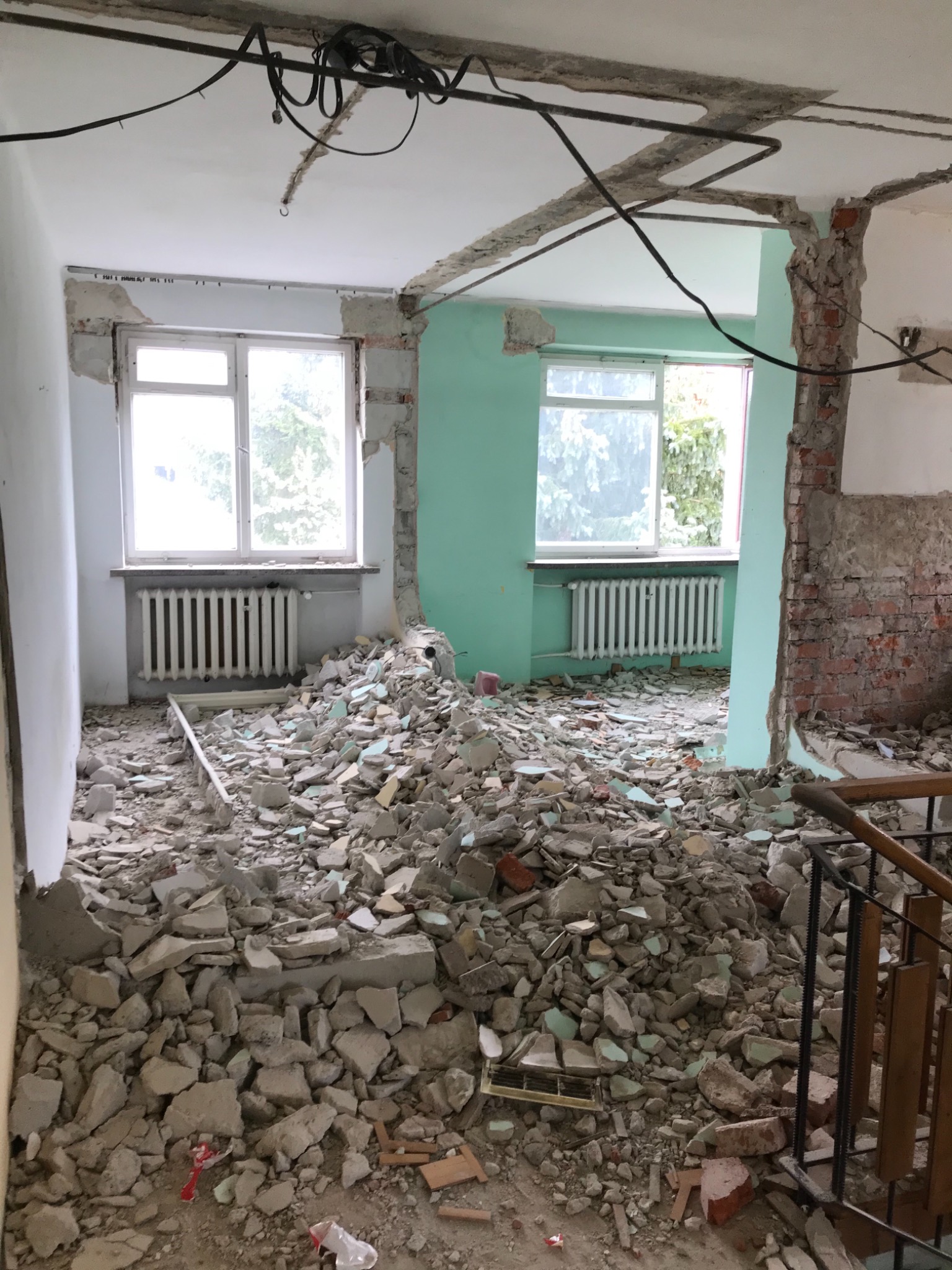 Wnętrze mieszkania w trakcie remontu; gruz i resztki tynku na podłodze, odsłonięte cegły, okna z grzejnikami, instalacje elektryczne wystające z sufitu.