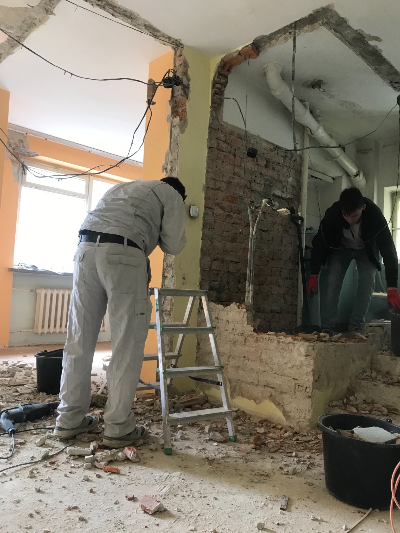 Dwóch mężczyzn w trakcie prac rozbiórkowych ściany działowej wewnątrz budynku, widoczne odsłonięte cegły, instalacje i gruz na podłodze, drabina oparta o ścianę.