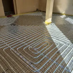 Białe rury ogrzewania podłogowego ułożone spiralnie na czarnej siatce, widoczne logo Keller, w tle surowy stan wykończenia wnętrza.