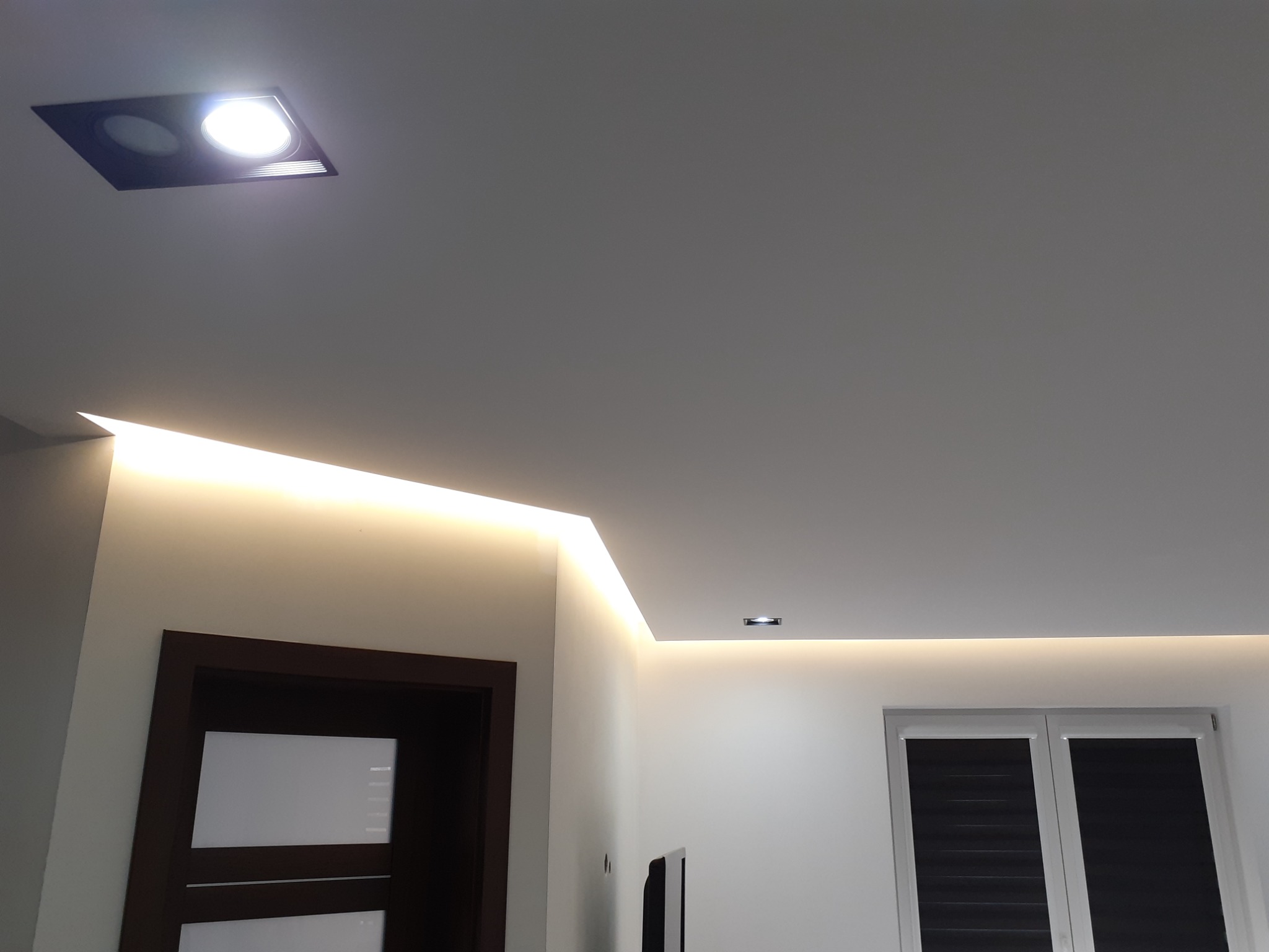 Nowoczesny sufit podwieszany z oświetleniem LED wzdłuż krawędzi, punktowe oświetlenie w suficie, widoczny fragment ściany z drzwiami w ciemnej ramie i oknem z żaluzjami.