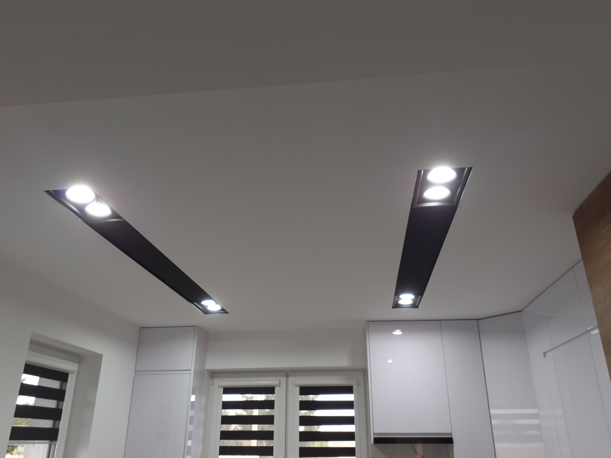 Biały sufit z dwoma podłużnymi wnękami z czarnym wykończeniem, w których zamontowano punktowe oświetlenie LED, widok na fragment białej zabudowy kuchennej i okno z roletami.