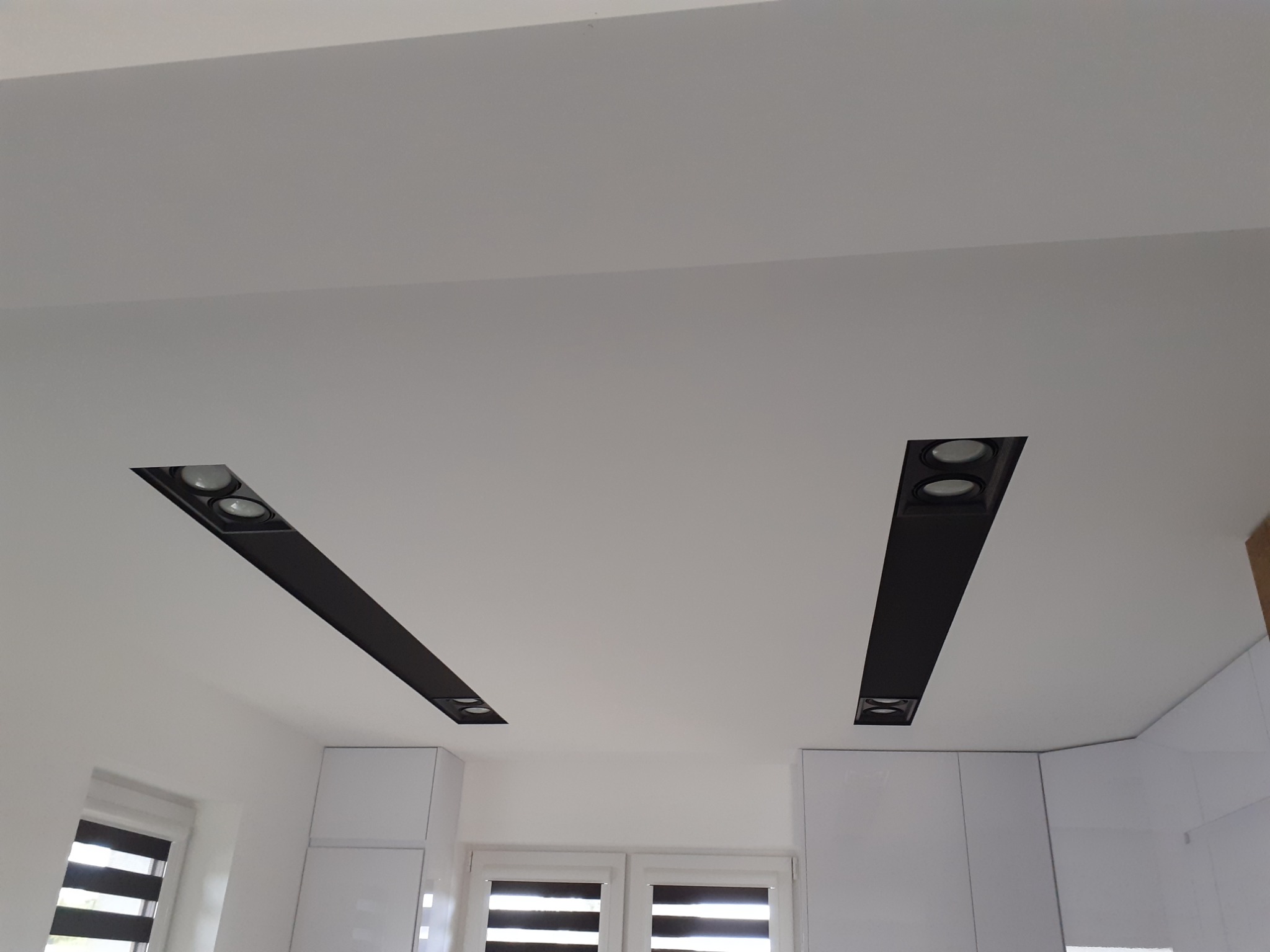 Biały sufit z dwoma podłużnymi, czarnymi oprawami oświetleniowymi z reflektorami, widoczne białe szafki kuchenne i okno z żaluzjami.