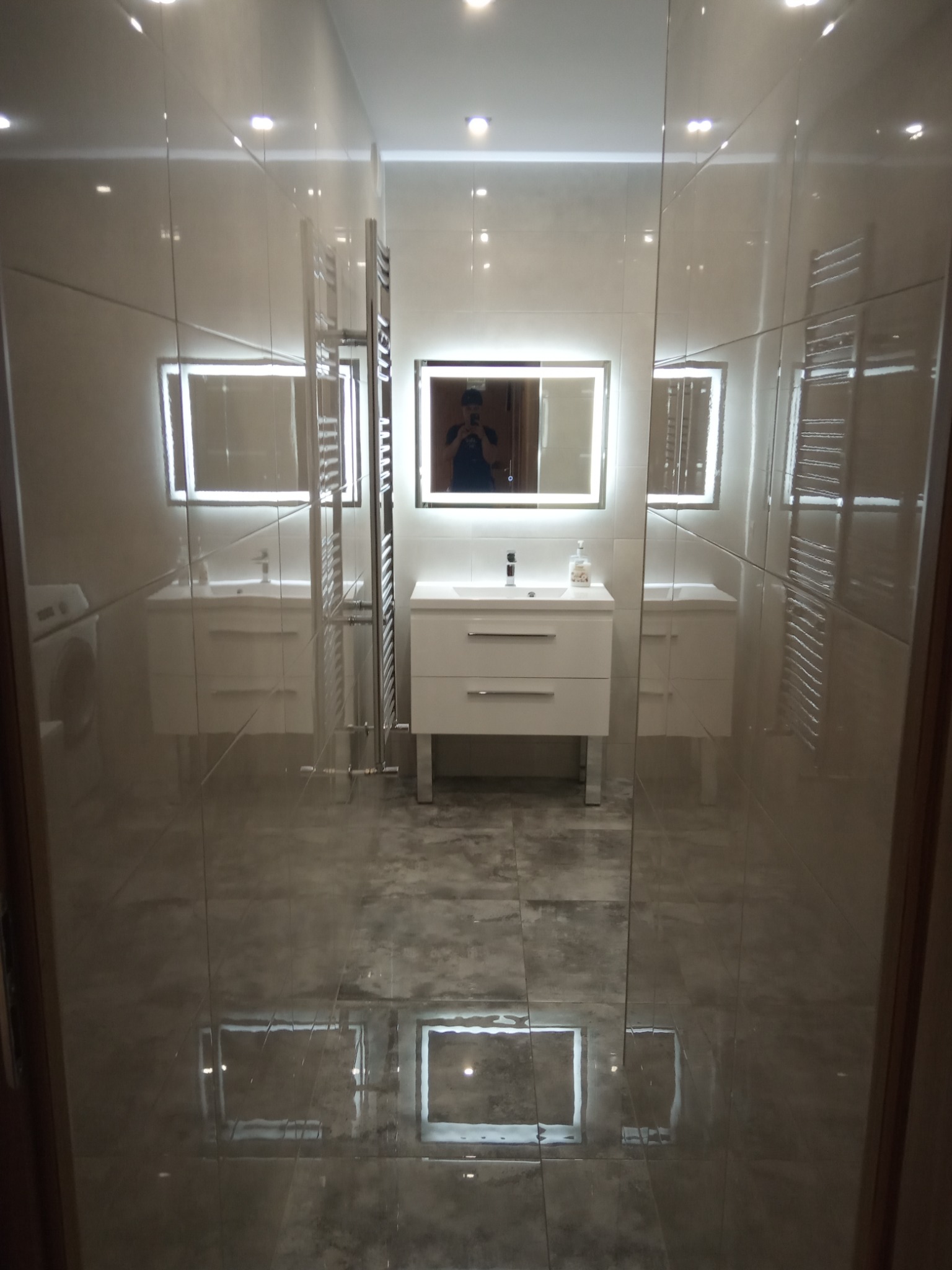 Nowoczesna łazienka z szarymi płytkami, podświetlanym lustrem nad białą szafką z umywalką oraz grzejnikiem drabinkowym, widoczna przez szklaną ścianę kabiny prysznicowej.