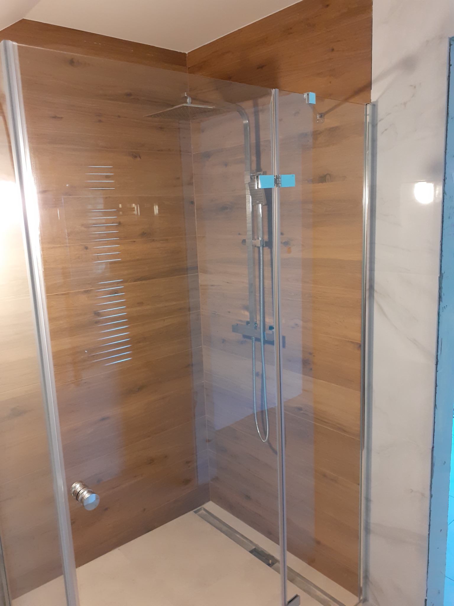 Kabina prysznicowa z deszczownicą, wykończona płytkami imitującymi drewno, z odpływem liniowym w podłodze, widok z boku.
