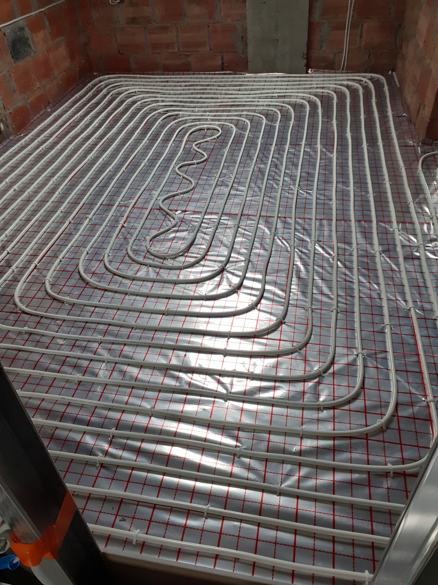 Białe rury ogrzewania podłogowego ułożone w pętle na srebrnej folii z czerwoną kratką, widok z góry, surowy mur z cegły w tle.