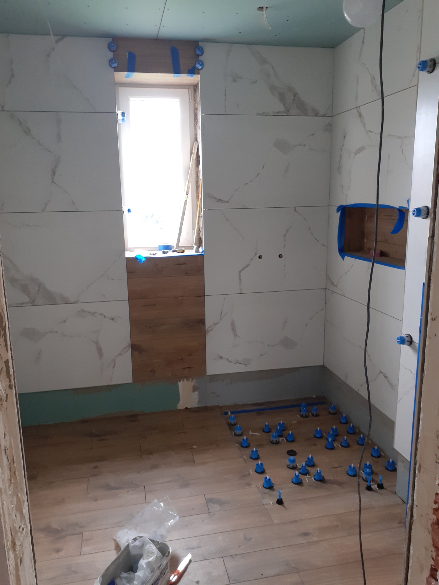 Wnętrze łazienki w trakcie prac w Złotej: układanie płytek imitujących marmur na ścianach oraz drewnopodobnych na podłodze, widoczne systemy poziomowania płytek i wnęka w ścianie przygotowana...