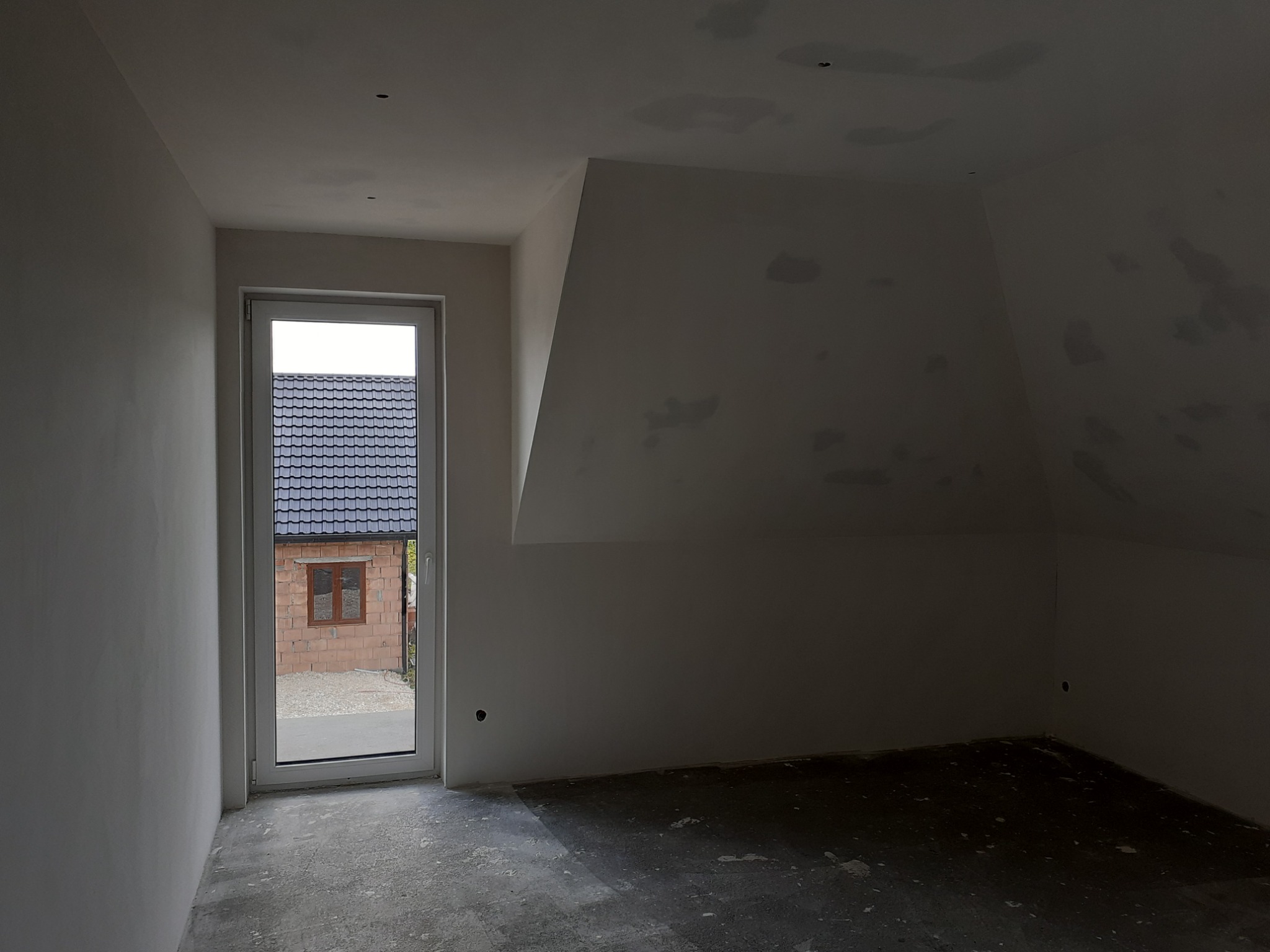 Poddasze w stanie deweloperskim z oknem, ściany i sufit zagruntowane, widoczne ślady po szpachlowaniu, surowa betonowa podłoga.