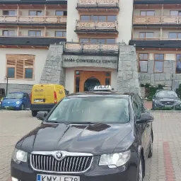 Czarny samochód taxi marki Skoda Superb z tablicą rejestracyjną KMY LE78 na tle Hotelu Bania Conference Center w Zakopanem, obok zaparkowane inne pojazdy.