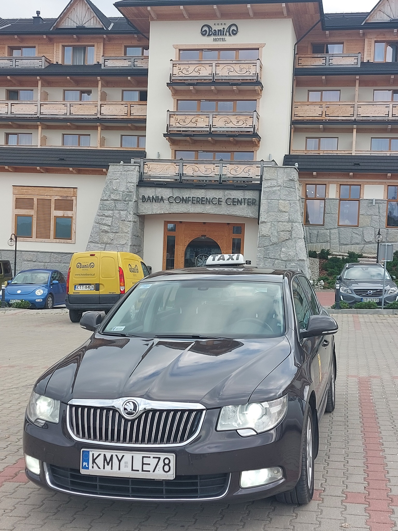 Czarny samochód taxi marki Skoda Superb z tablicą rejestracyjną KMY LE78 na tle Hotelu Bania Conference Center w Zakopanem, obok zaparkowane inne pojazdy.