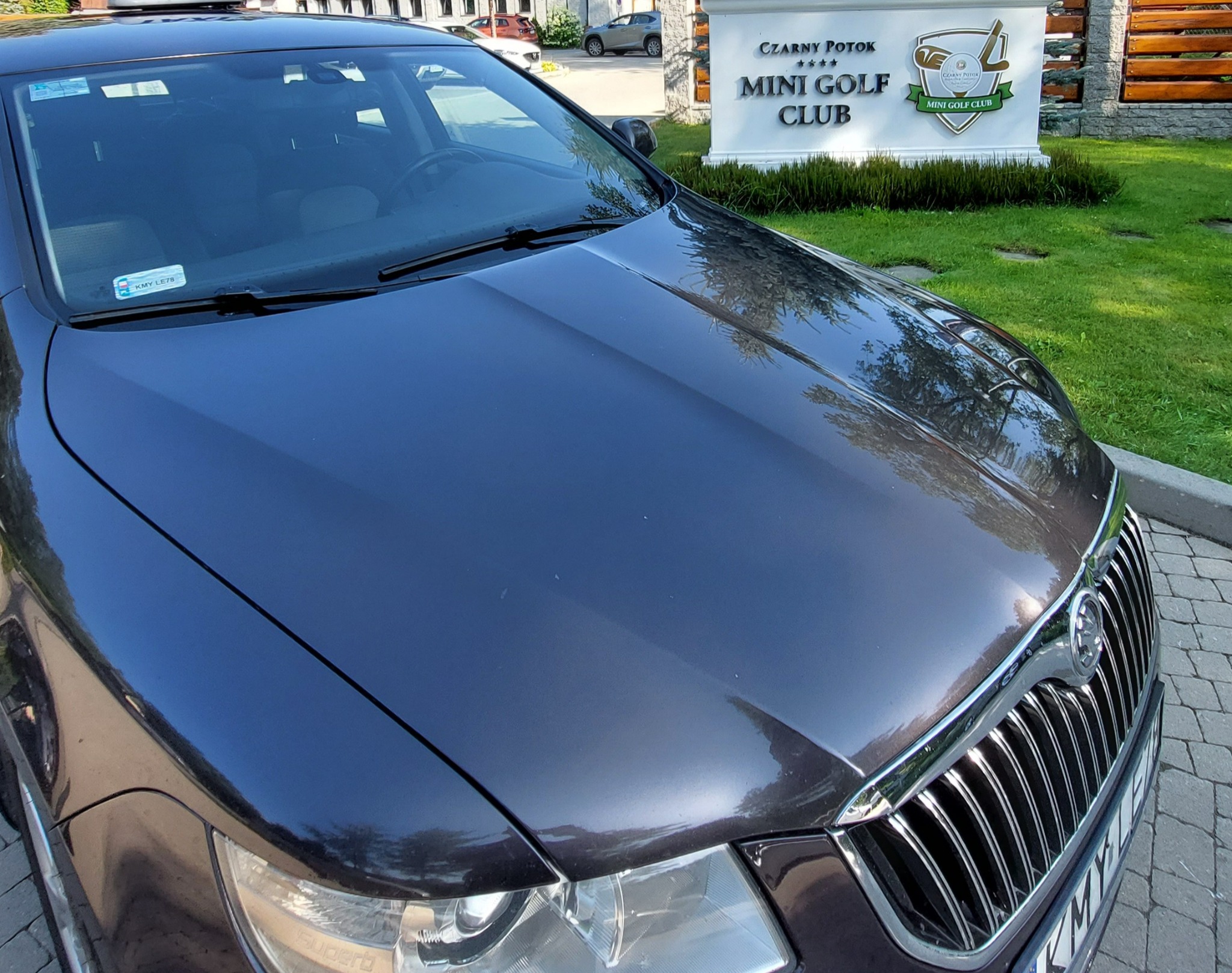 Ciemnoszary samochód Skoda Superb zaparkowany przed wejściem do Czarny Potok Mini Golf Club, widoczna tablica rejestracyjna KMY LE78.