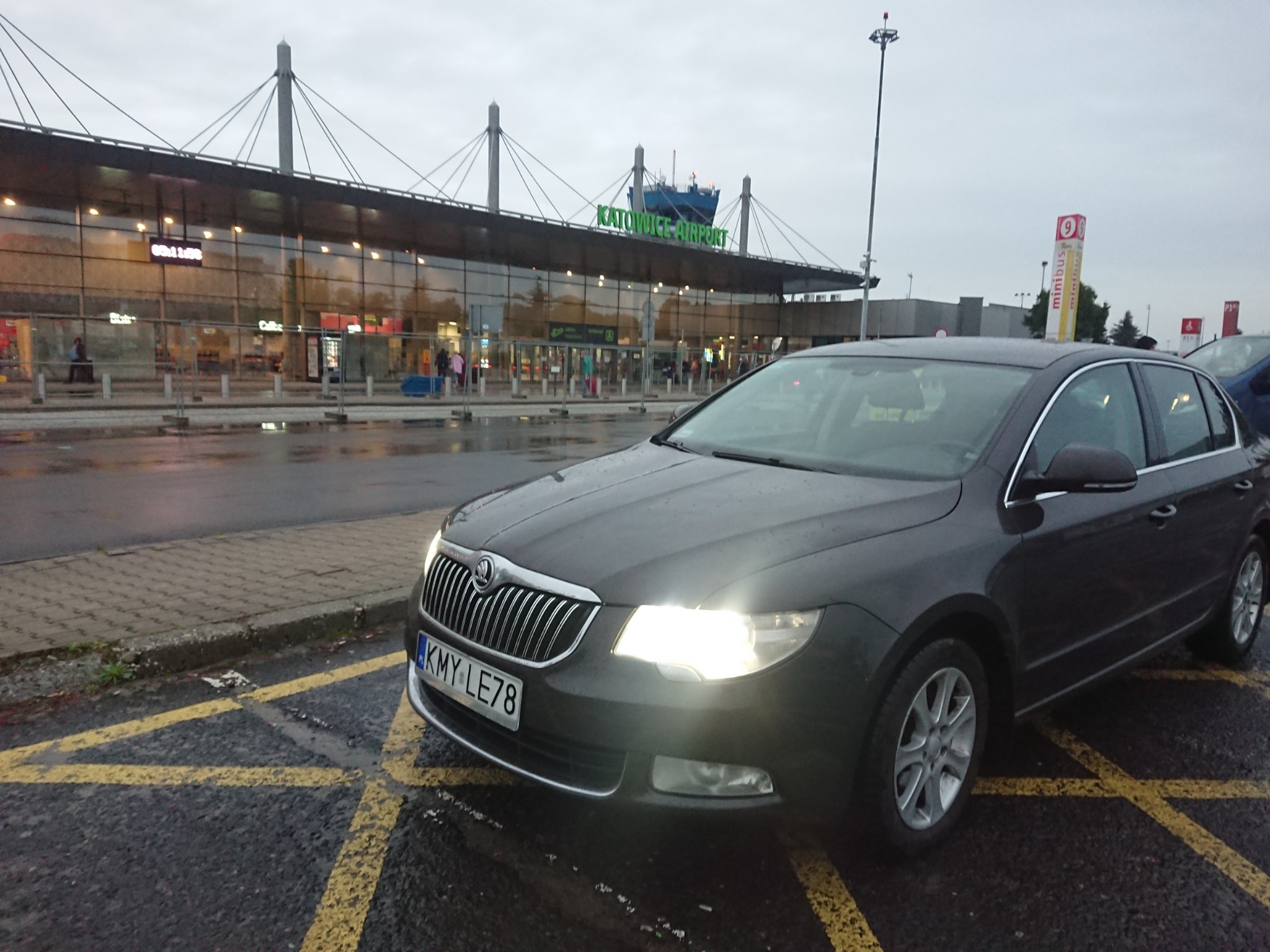 Ciemnoszara Skoda Superb z włączonymi światłami na parkingu lotniska Katowice-Pyrzowice w pochmurny dzień.