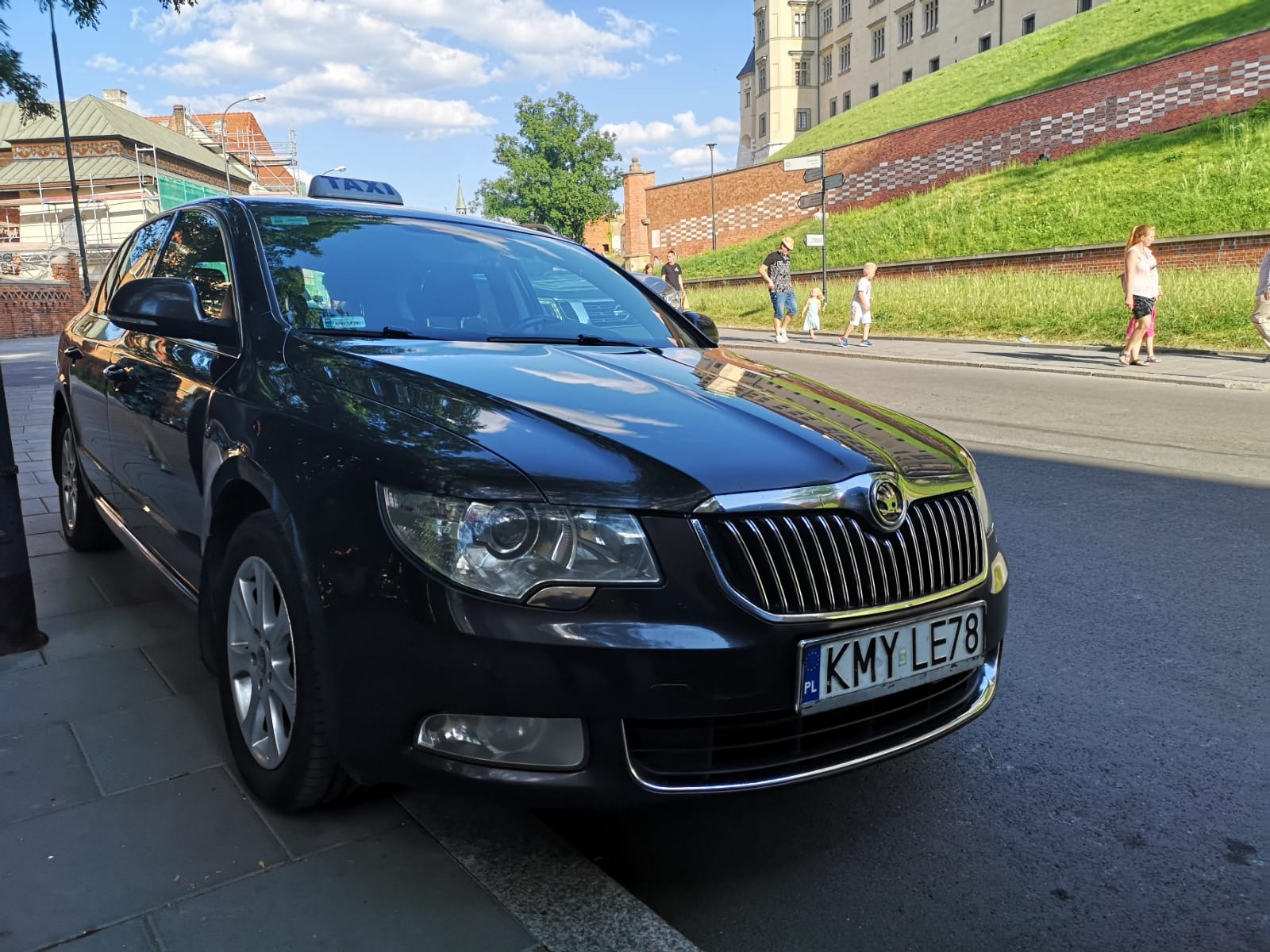 Czarna taksówka Skoda Superb z numerem rejestracyjnym KMY LE78, zaparkowana na ulicy z widokiem na Wawel w Krakowie.