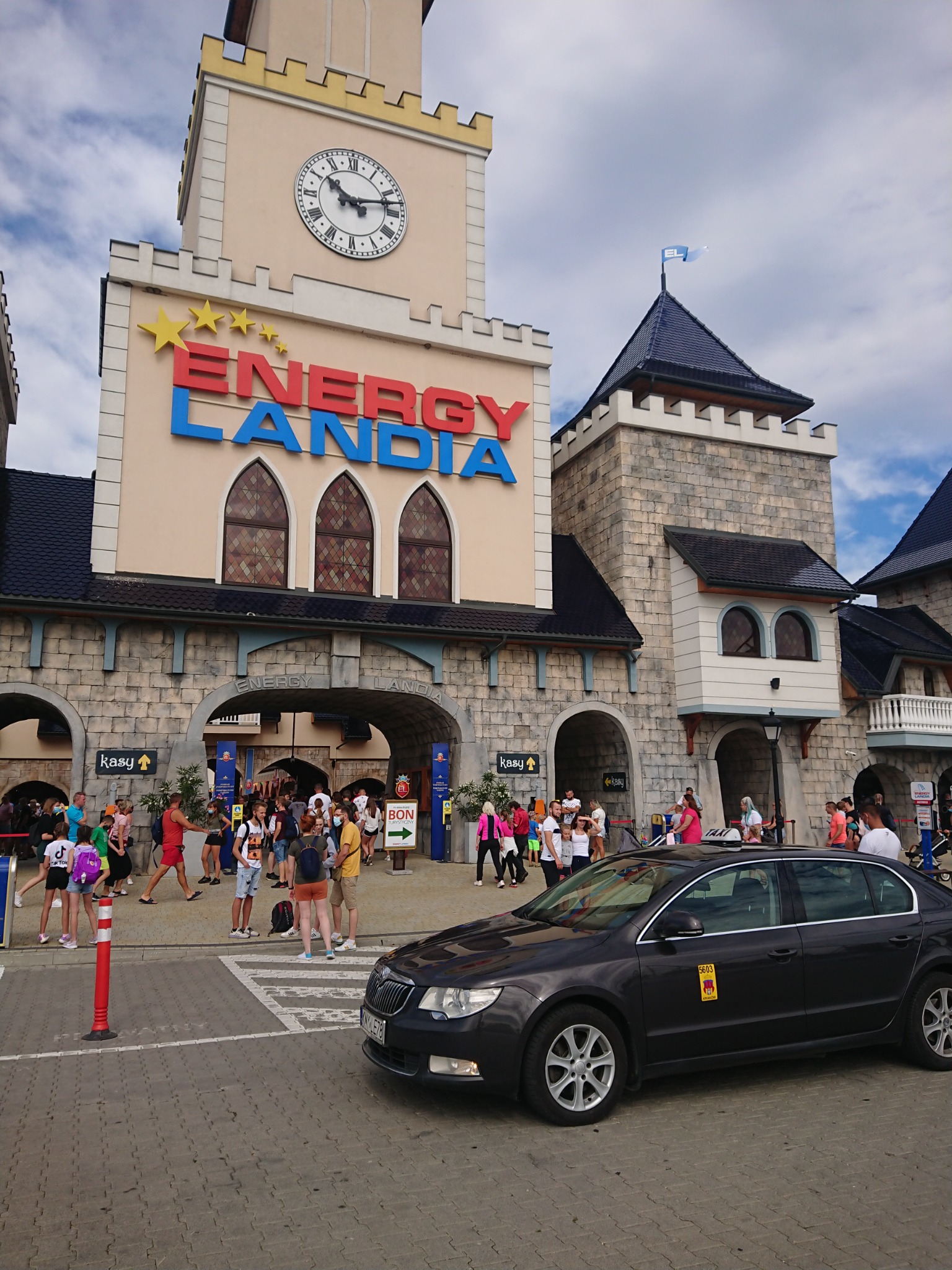 Ciemny samochód taxi z żółtym oznaczeniem na tle wejścia do parku rozrywki Energylandia z tłumem ludzi.