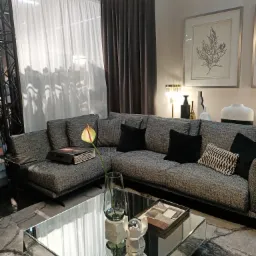 Szara narożna sofa z czarnymi poduszkami i wzorzystą poduszką, na szklanym stoliku kawowym dekoracje, w tle białe i grafitowe zasłony, oświetlenie sufitowe i grafika na ścianie.