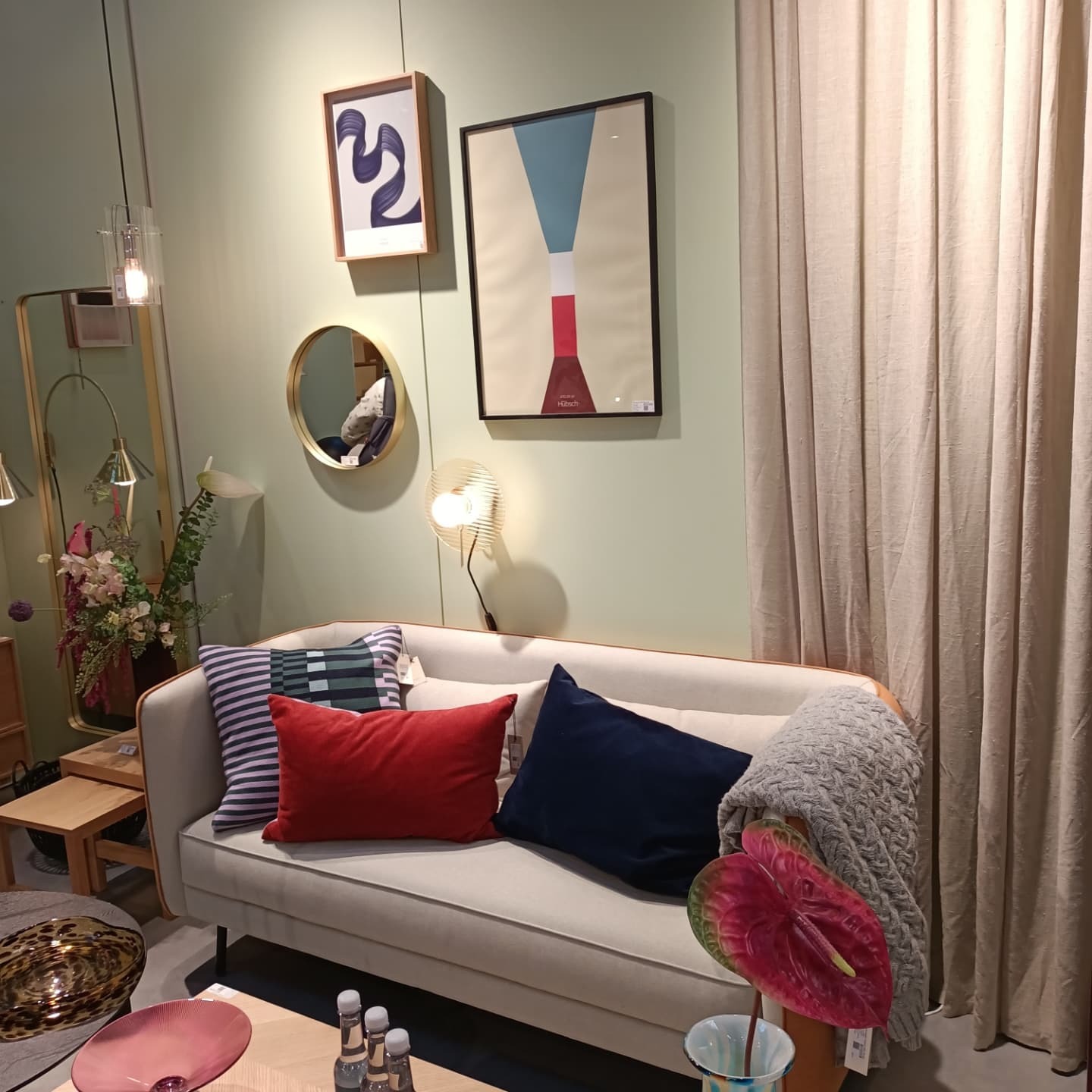 Jasna sofa z dekoracyjnymi poduszkami i pledem, uzupełniona o kwiat anturium w wazonie, stolik kawowy z szklanymi butelkami wody, na tle ściany z obrazami i lustrem.