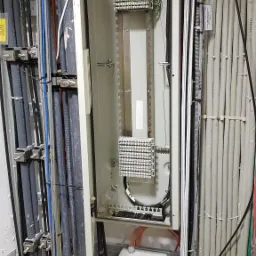 Panel krosowniczy z kablami telekomunikacyjnymi w szafie, widoczne złącza i przewody światłowodowe, skrzynka z napisem 'UWAGA Kabel Światłowodowy'.