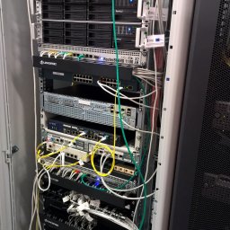 IPv6 Marta Musiał - Szafa serwerowa z zainstalowanymi urządzeniami sieciowymi różnych producentów: Dell, Synology, Juniper, MikroTik, Linkbasic, z widocznymi kablami połączeniowymi w kolorach białym, zielonym i żółtym.