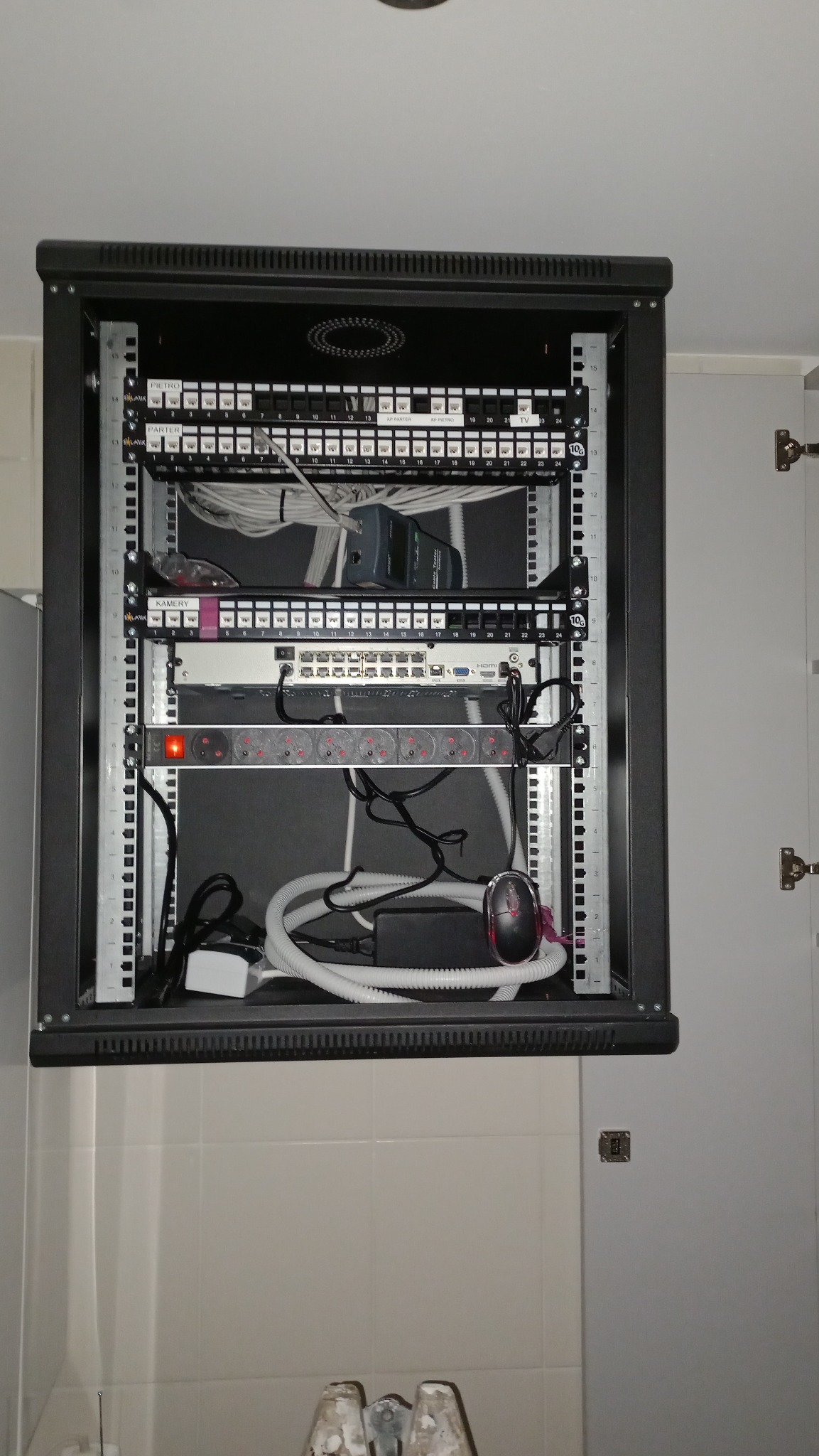 Szafa rack z patch panelem, switchem, listwą zasilającą i okablowaniem, umieszczona w szafce w pomieszczeniu z białymi płytkami.