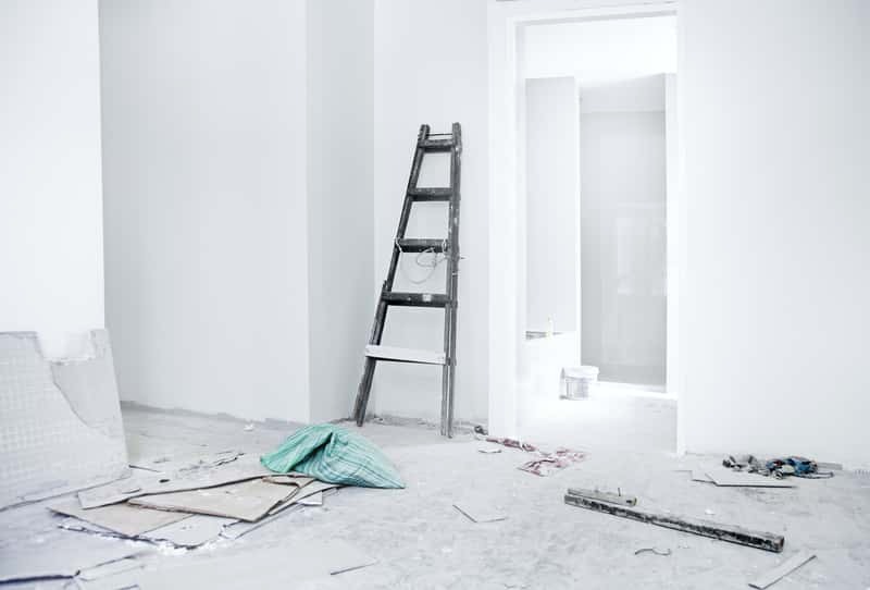 Pokój w trakcie prac przygotowawczych: białe ściany po tynkowaniu, drewniana drabina oparta o ścianę, zabezpieczenia podłogi z kartonu, torba z materiałami budowlanymi i widok na jasne wnętrze...