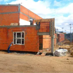 Budowa domu jednorodzinnego w stanie surowym otwartym: ściany z czerwonej cegły, wstawione okna PCV, widoczne zbrojenia i szalunki, teren budowy.